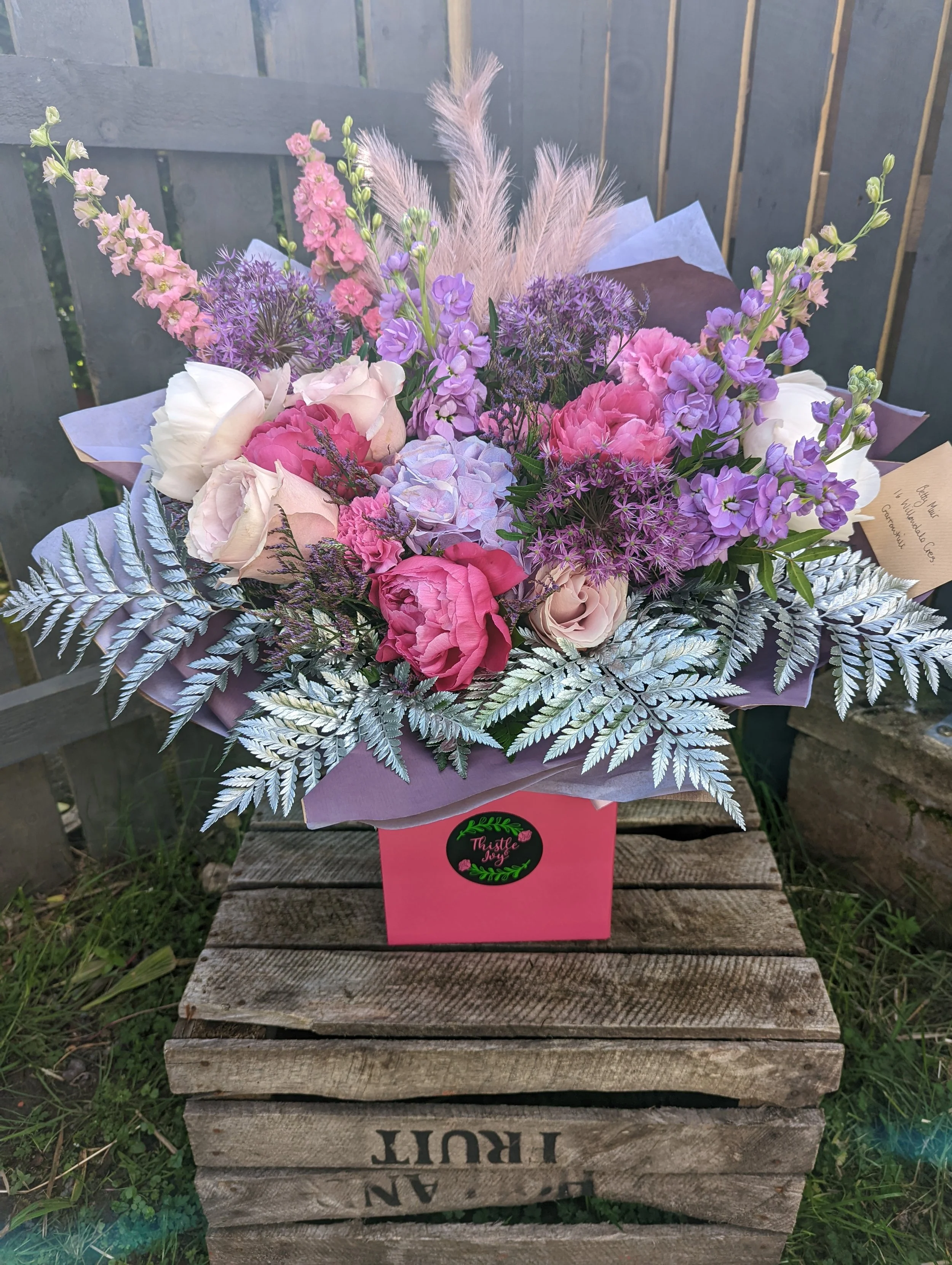 A colorful bouquet of pink, purple, cream, and white flowers, wrapped in purple tissue paper, in a pink box with a