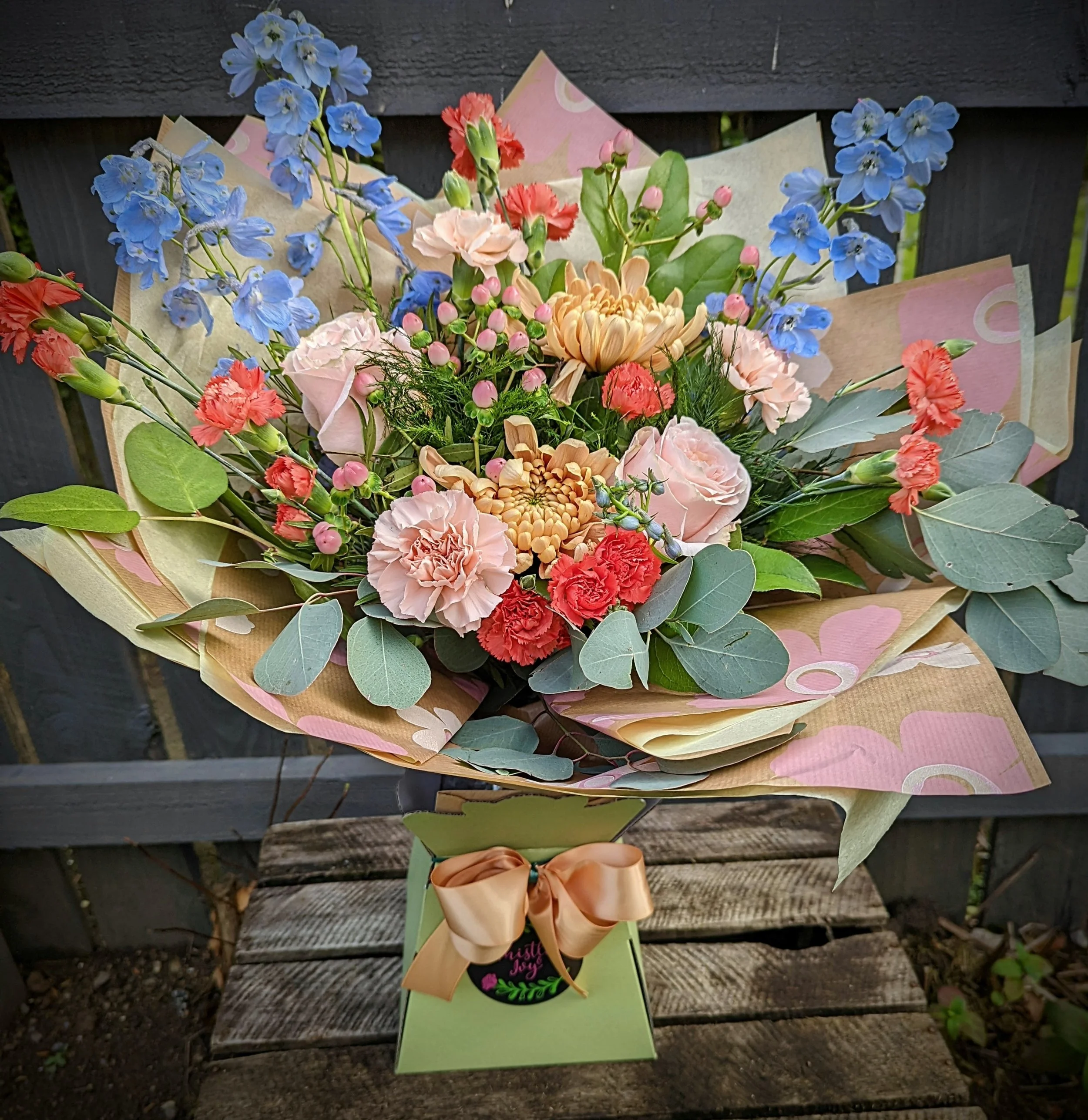 A colorful bouquet of mixed flowers, including pink roses, blue delphiniums, peach chrysanthemums, red and pink carnations, and pink hypericum berries, arranged with green leaves and wrapped in patterned paper, tied with a beige satin bow, and placed on a green stand with a chalkboard label reading 'Just for you'.