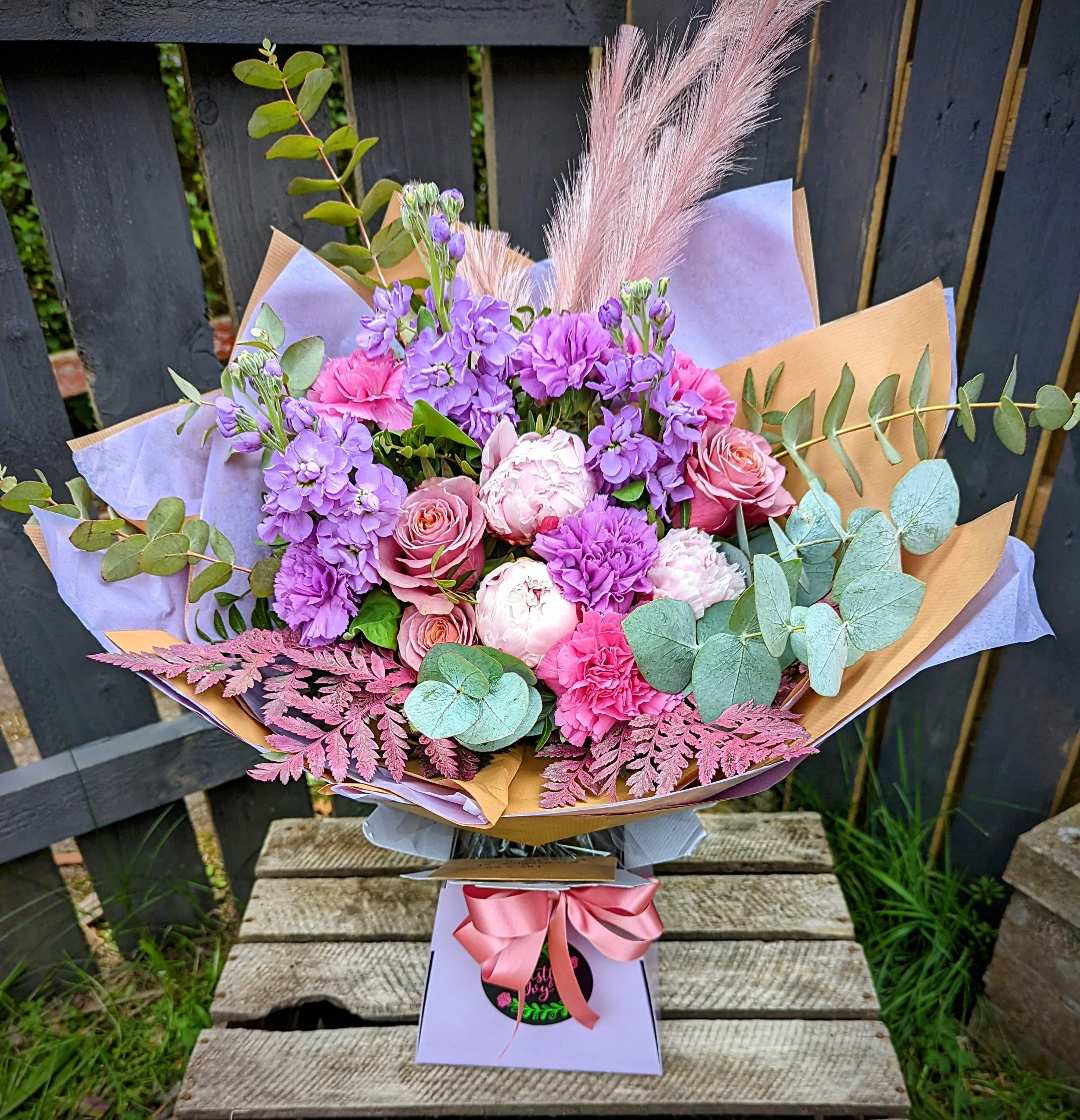 A colorful bouquet of pink, purple, and white flowers with green foliage, pink feathery accents, wrapped in tan and purple paper, with a pink ribbon, placed on a wooden bench outdoors.
