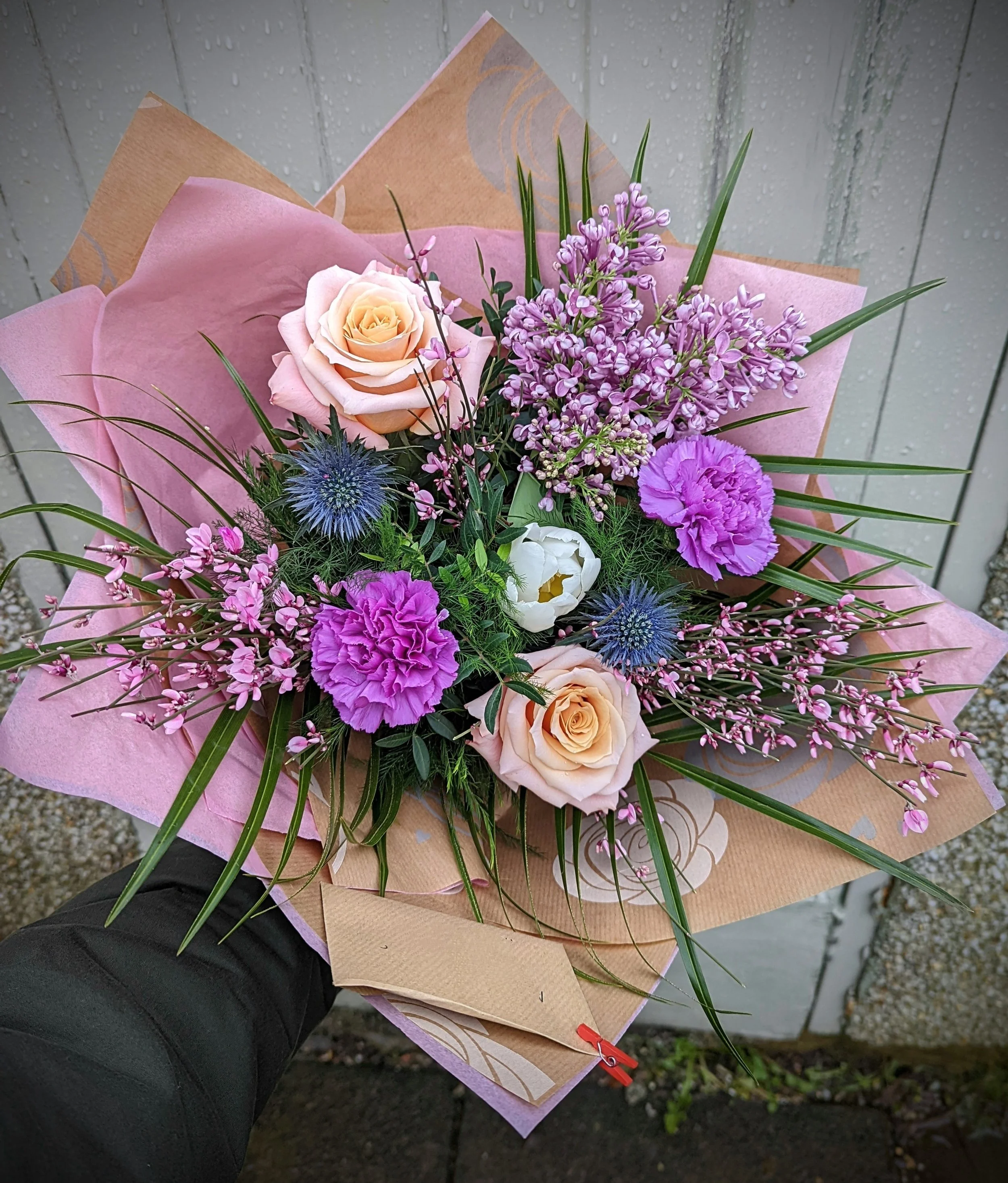 A colorful bouquet of flowers including pink roses, purple carnations, white lisianthus, blue sea holly, and pink wax flowers, wrapped in pink and brown paper, being held outdoors against a gray wooden background.