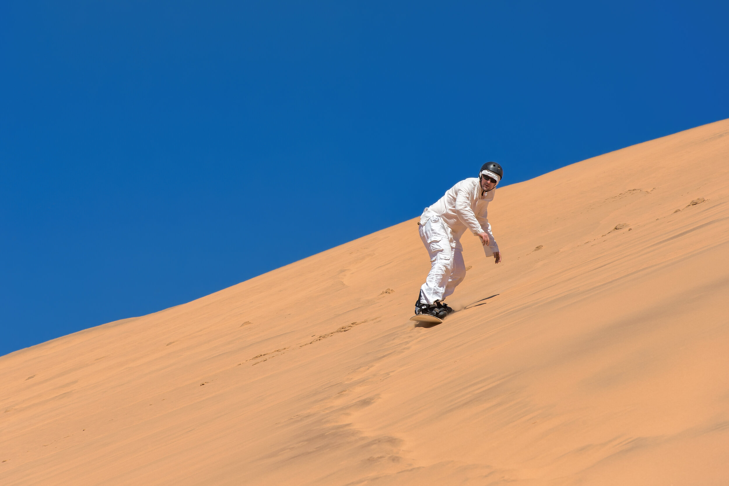 man wearing white bodysuit sandboarding down sand dune