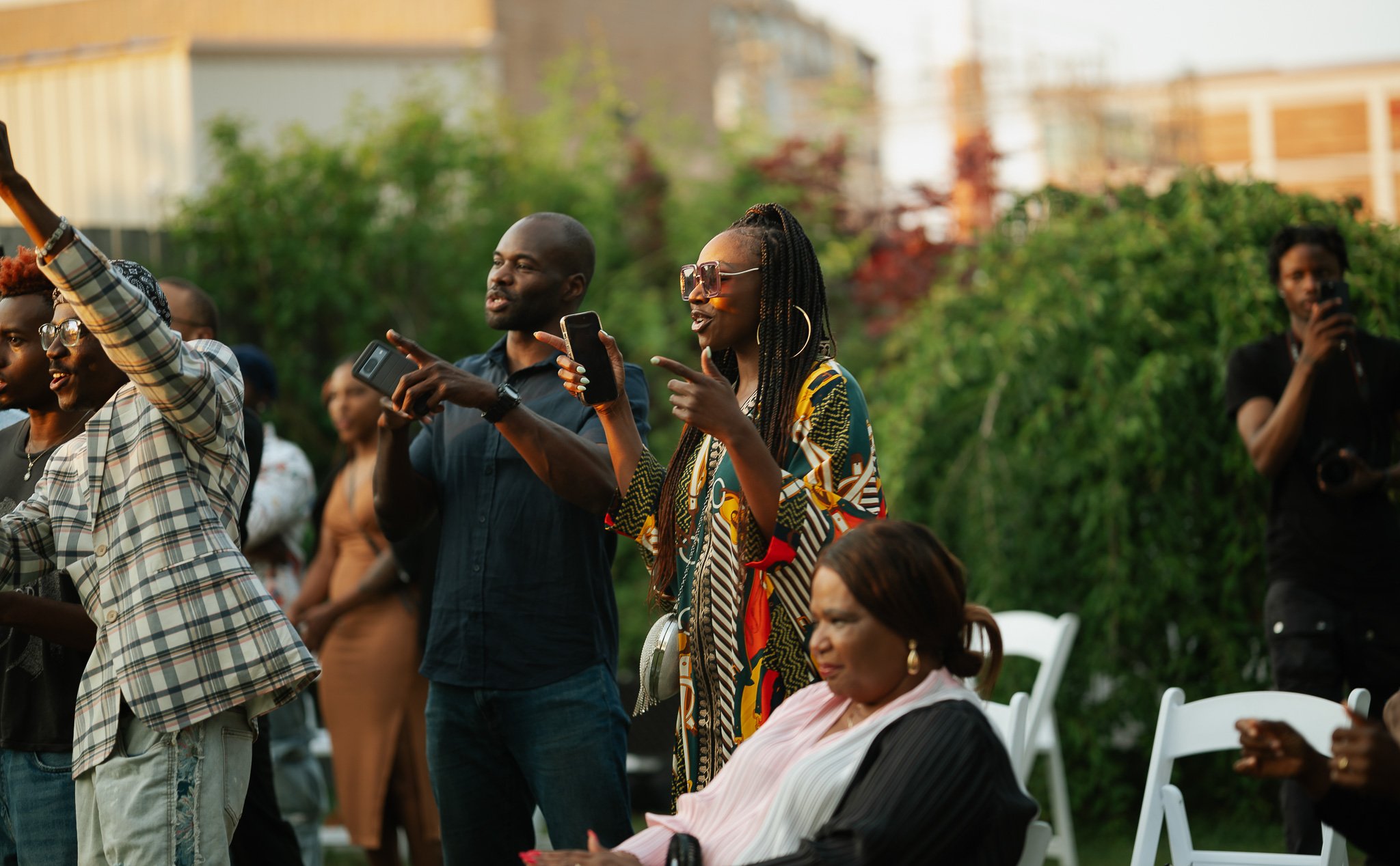 Group of people outdoors, some standing and some seated, enjoying an event during daylight. The background features greenery and blurred buildings.