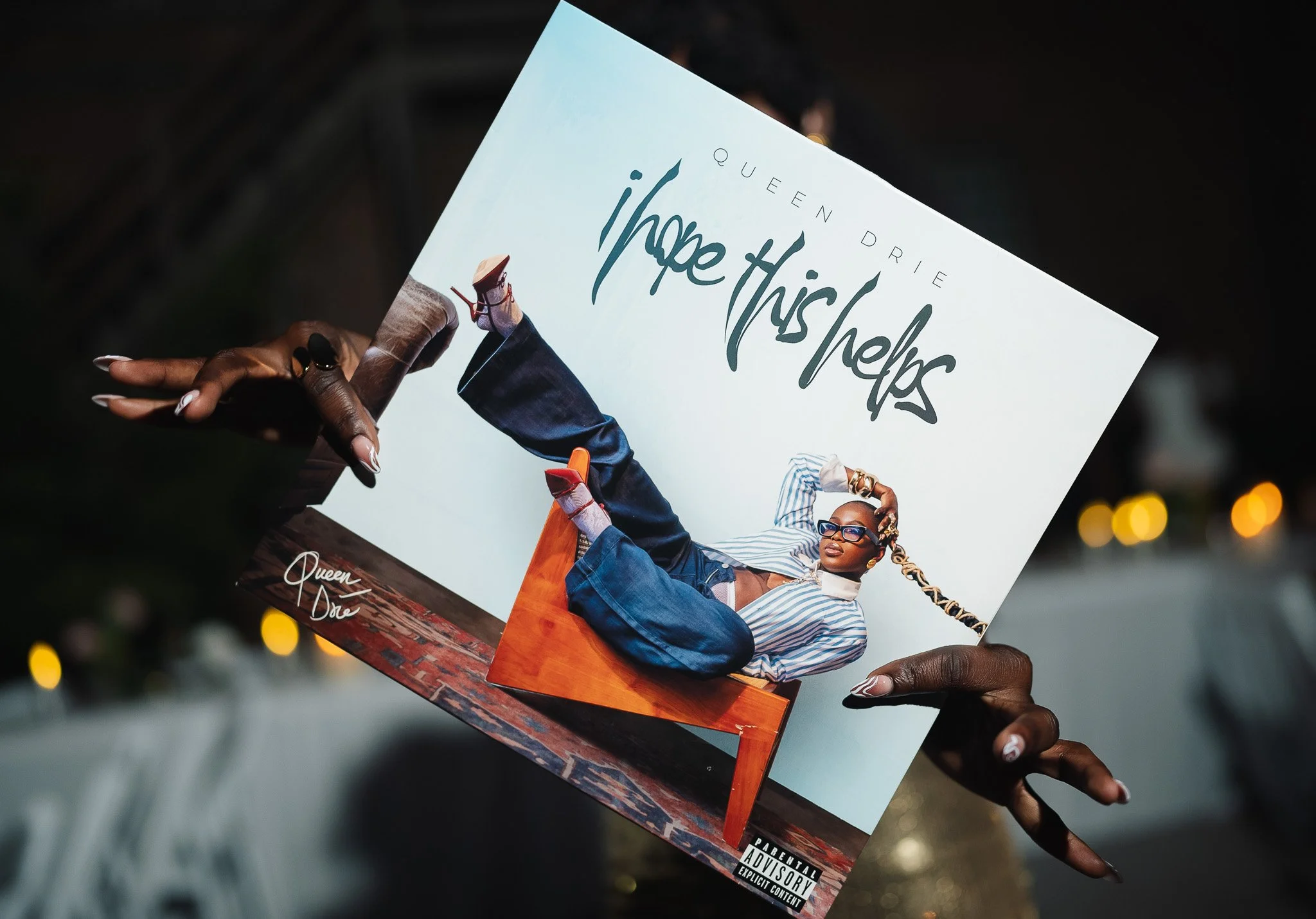 Album cover for Queen Drie's 'i hope this helps', featuring a woman with braided hair, glasses, and stylish clothing, sitting on a wooden table with a light blue background.