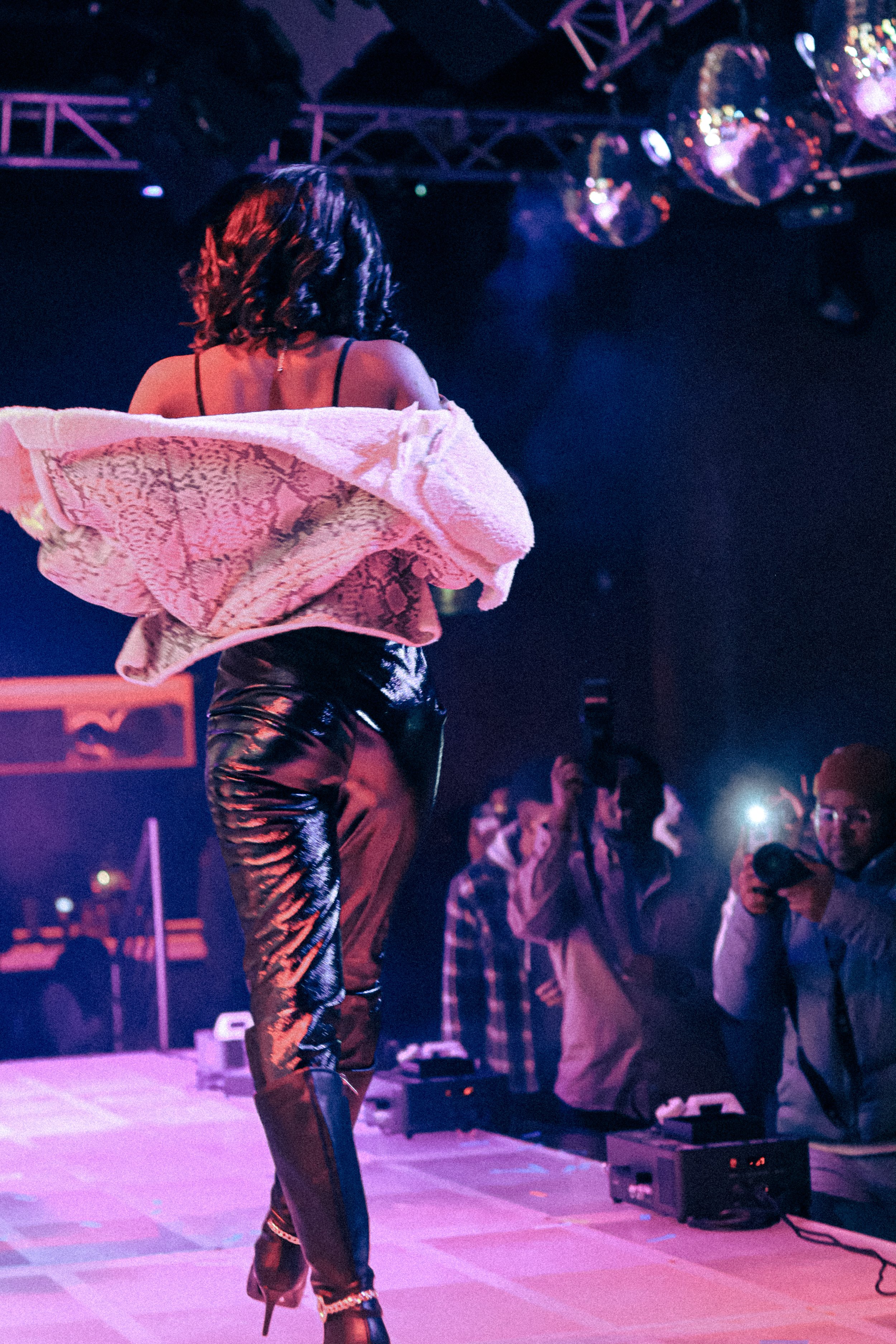 A woman performing on stage in a nightclub, wearing shiny pants, a black top with thin straps, and a pink textured jacket draped over her shoulders. Several audience members watch and take photos, with stage lights and disco balls overhead.