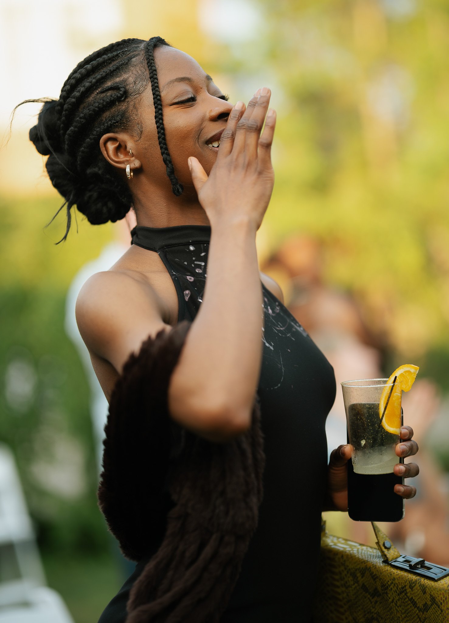 A woman with braided hair wearing a black dress and a fur coat draped over her shoulders is holding a glass of sparkling drink garnished with a lemon wedge and a straw, while smiling and appears to be enjoying herself outdoors.