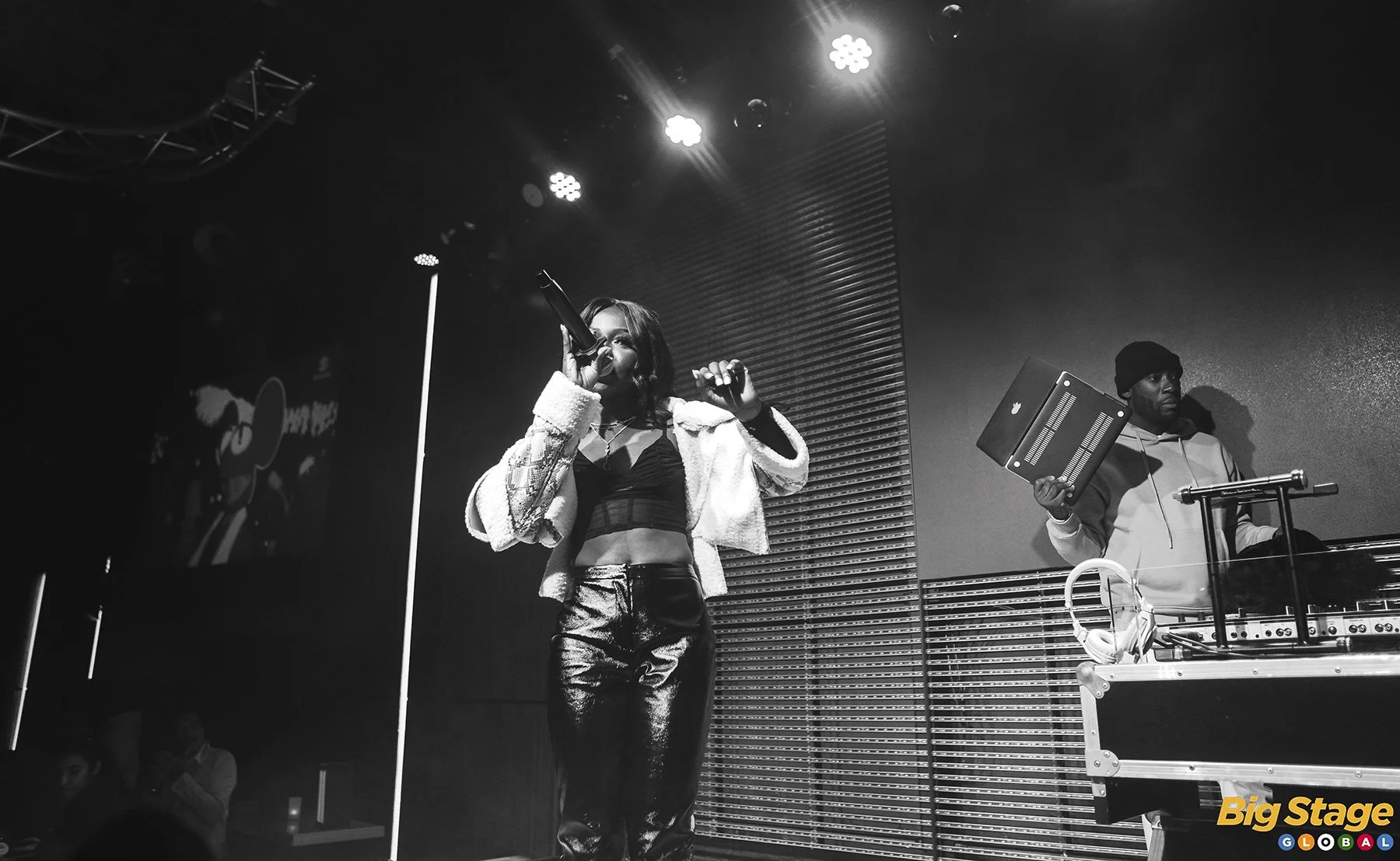 A female singer with shoulder-length dark hair in a black crop top and shiny pants performs on stage with a microphone, accompanied by a DJ in a hoodie and beanie manipulating music equipment. Stage lighting is visible overhead.