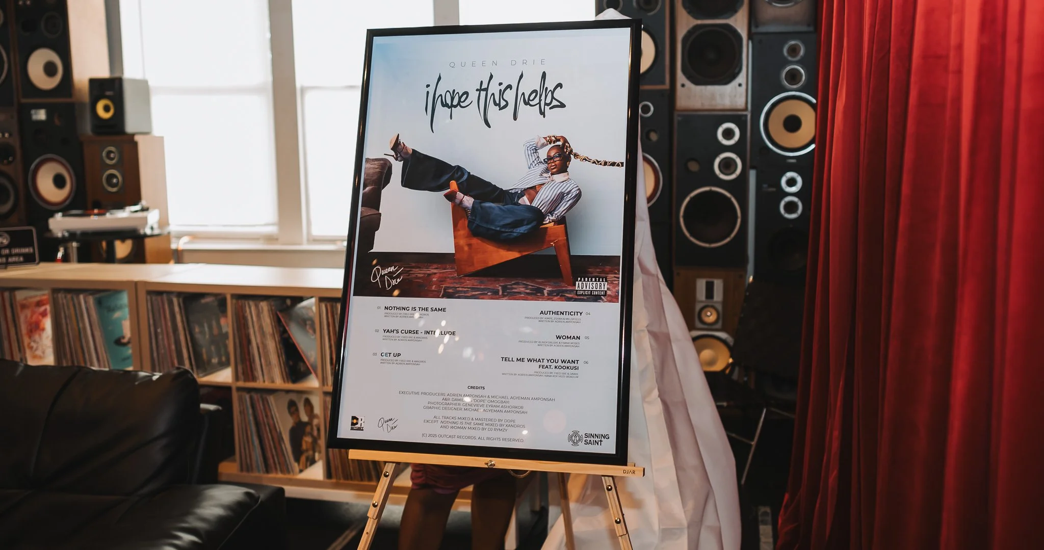 A promotional poster for Queen Drie's music album titled "i hope this helps" displayed on an easel in a studio or event space with vinyl records, speakers, and red curtains in the background.