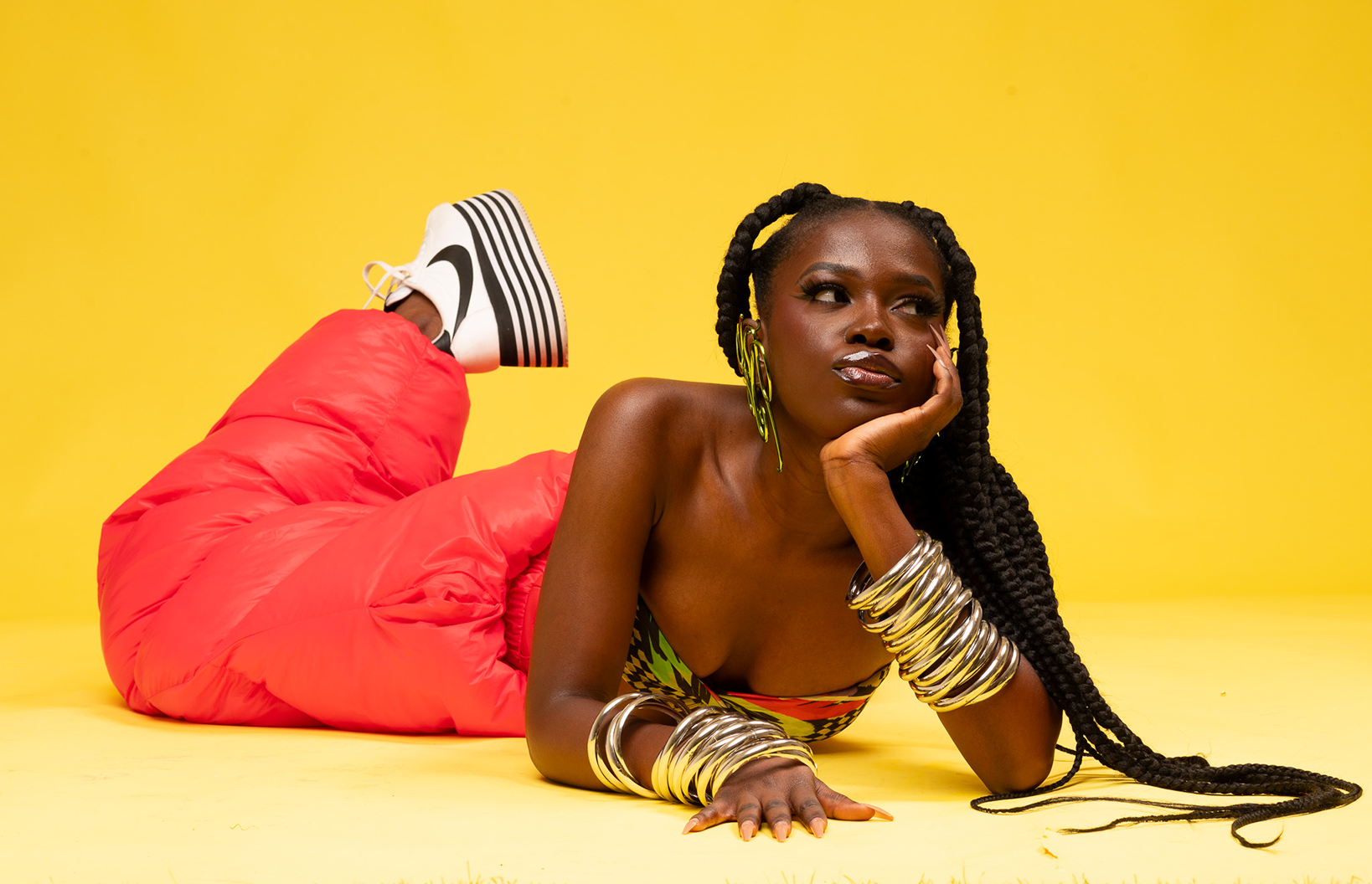Queen Drie with long braided hair, wearing colorful top, pink puffy pants, and black and white platform sneakers, lying on a yellow background, resting her head on her hand and looking to the side.