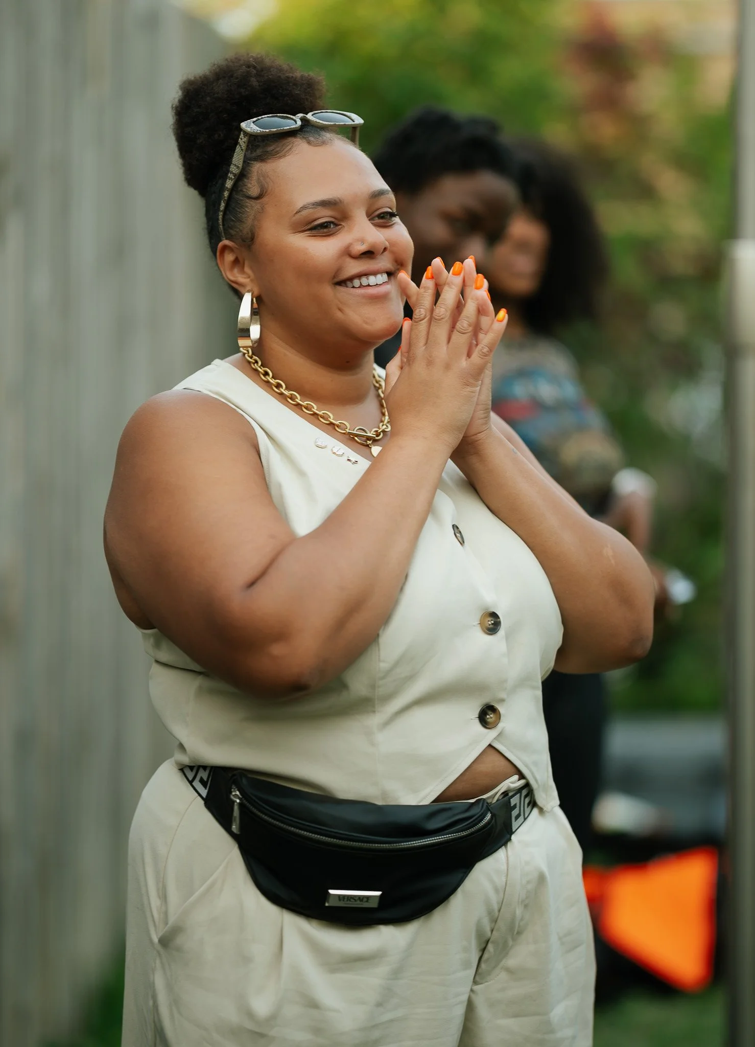 Smiling woman with sunglasses on her head, gold jewelry, and a fanny pack, standing outdoors with her hands clasped together, in a praying or hopeful gesture.