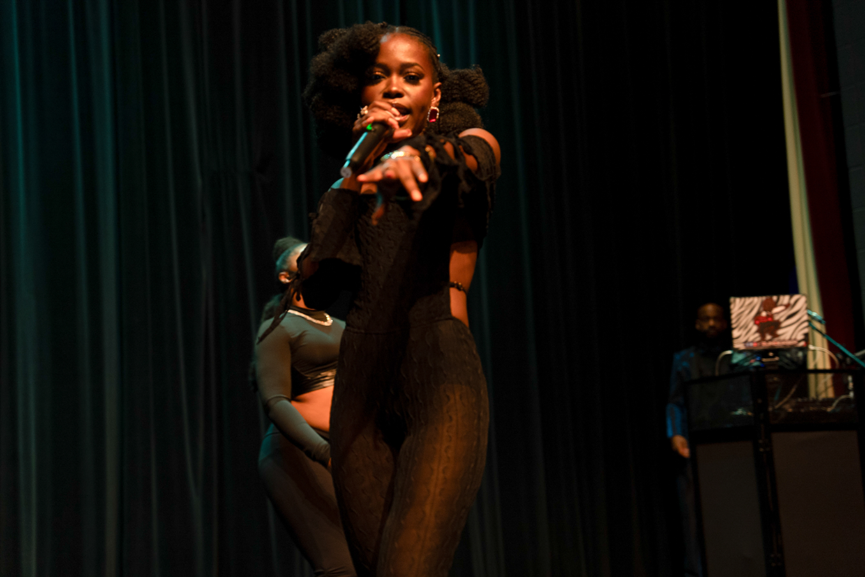 Queen Drie with curly hair singing into a microphone on stage, with a woman in the background and a man standing near a DJ booth.