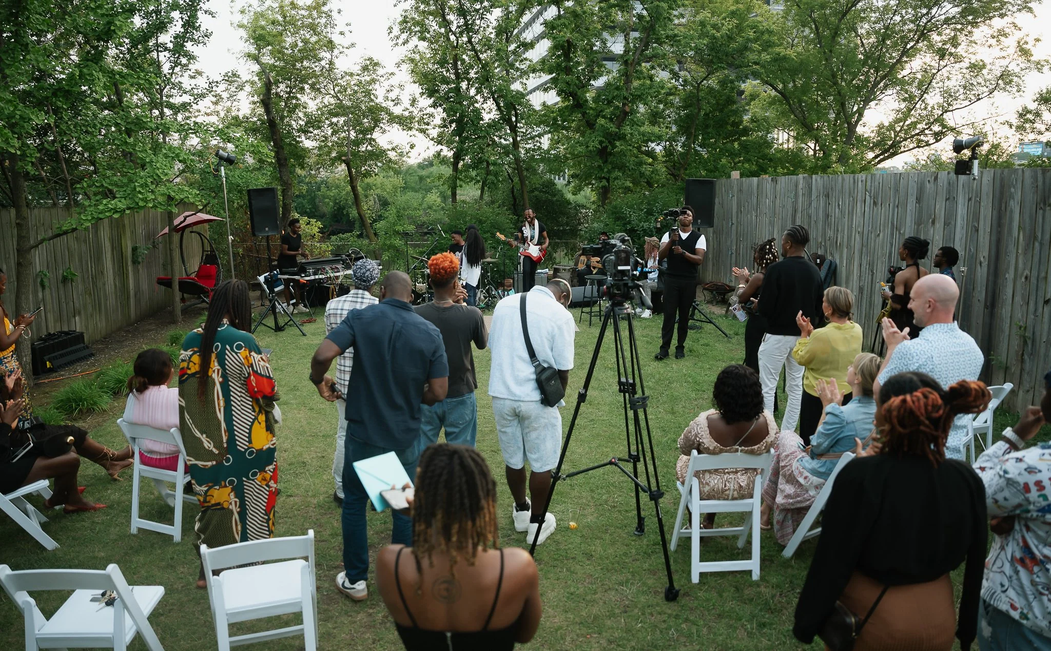 A diverse group of people attending an outdoor concert or event in a backyard, with some seated and others standing, watching musicians perform on a small stage surrounded by trees and a wooden fence.