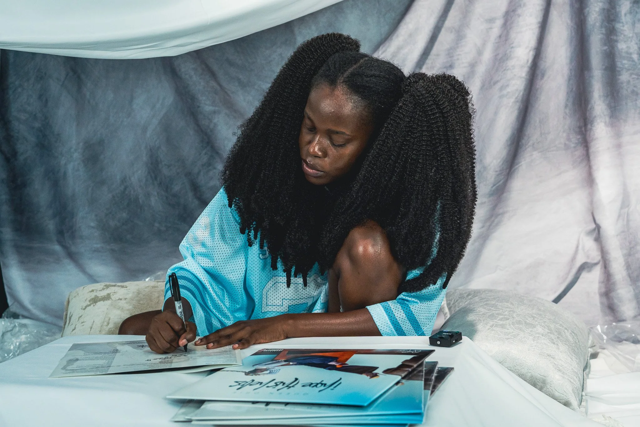 Queen Drie with long curly black hair, wearing a blue sports jersey, is sitting on a bed and signing vinyl copies of the EP 'I Hope This Helps'.