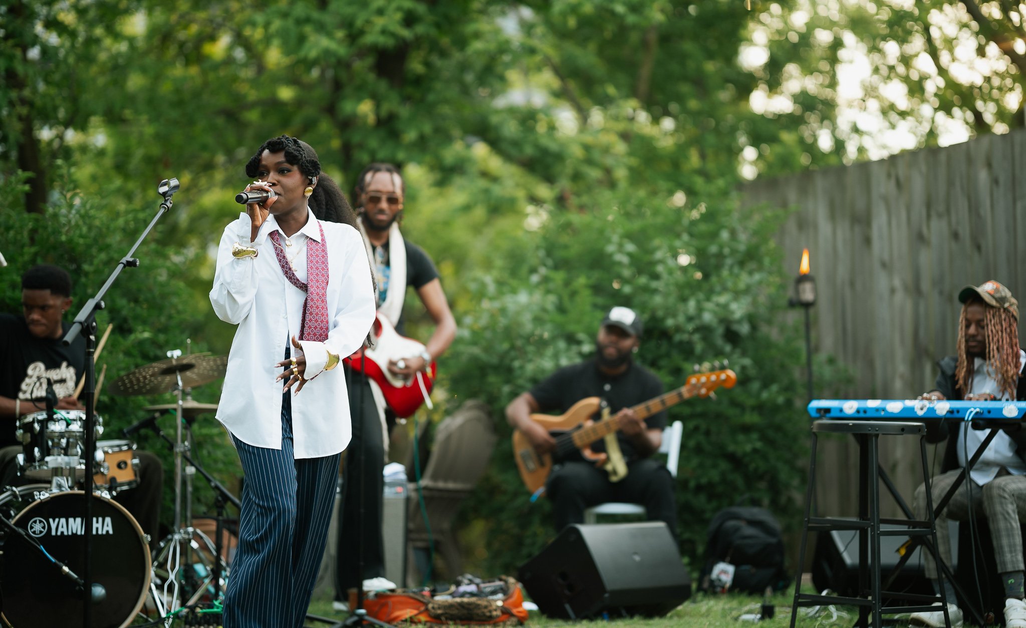 Queen Drie singing into a microphone with a band playing instruments behind her outdoors in a garden setting.