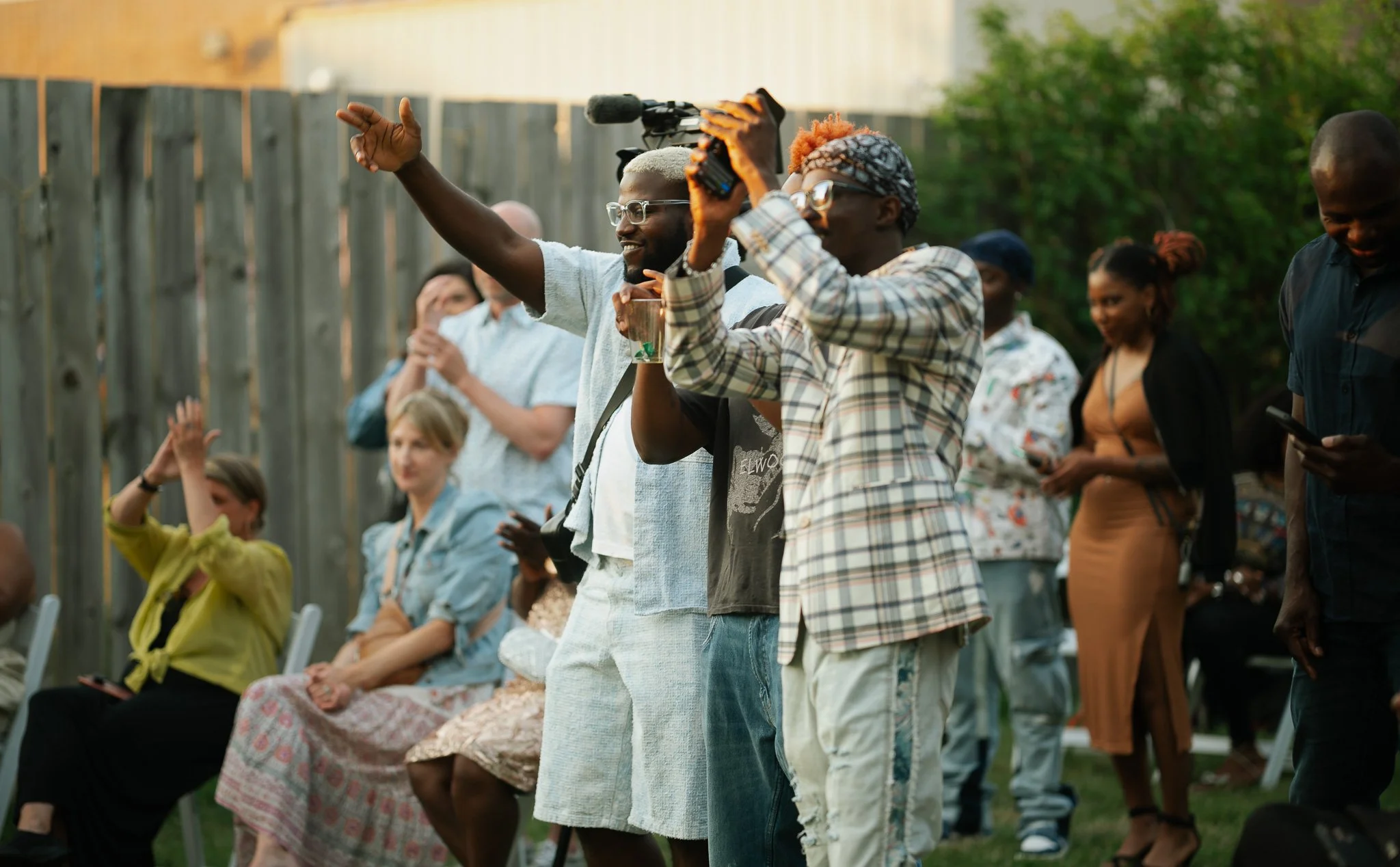 Group of people at an outdoor gathering, some standing and some sitting, with a wooden fence and greenery in the background. Most are engaged, some clapping or holding phones, enjoying the event.