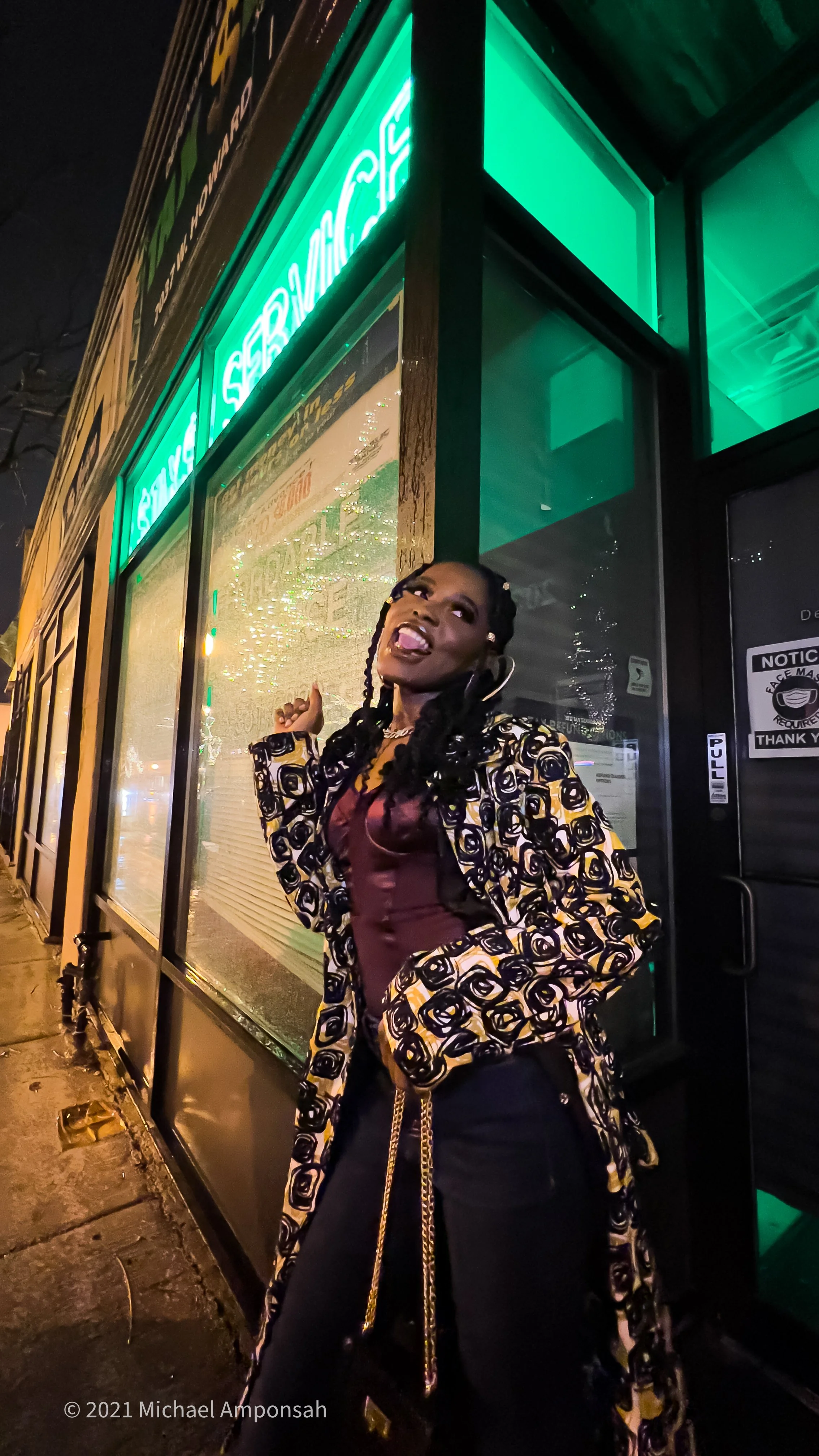 A woman stands outside a restaurant at night, making a playful face with her tongue sticking out, wearing a patterned coat and dark pants, illuminated by neon lights.