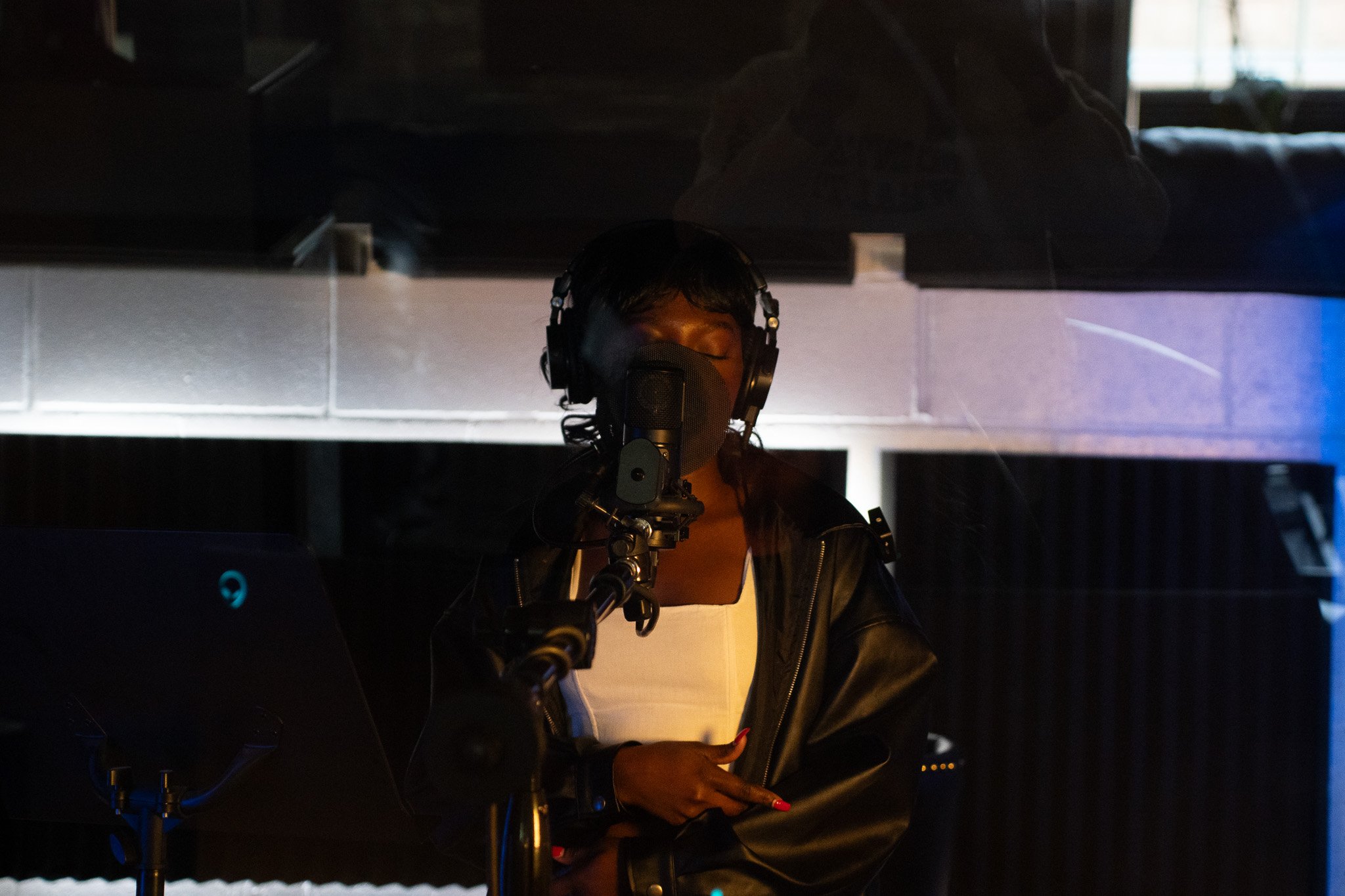 Queen Drie recording vocals in a sound studio, wearing headphones and a face mask, standing in front of a microphone, with music equipment and a computer nearby.