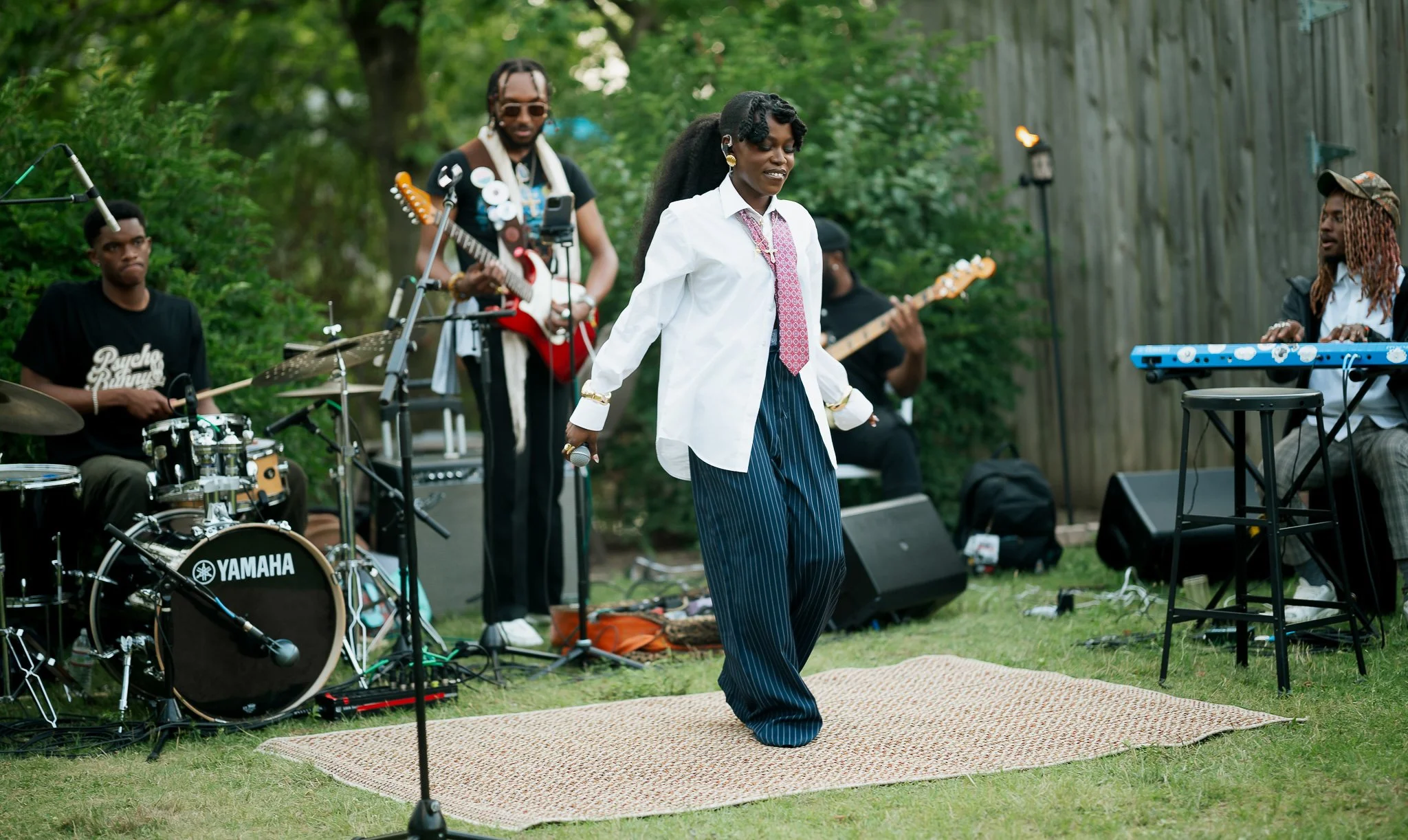 Queen Drie in a white shirt and striped pants dancing on a small rug while a live band performs outdoors in a green backyard.