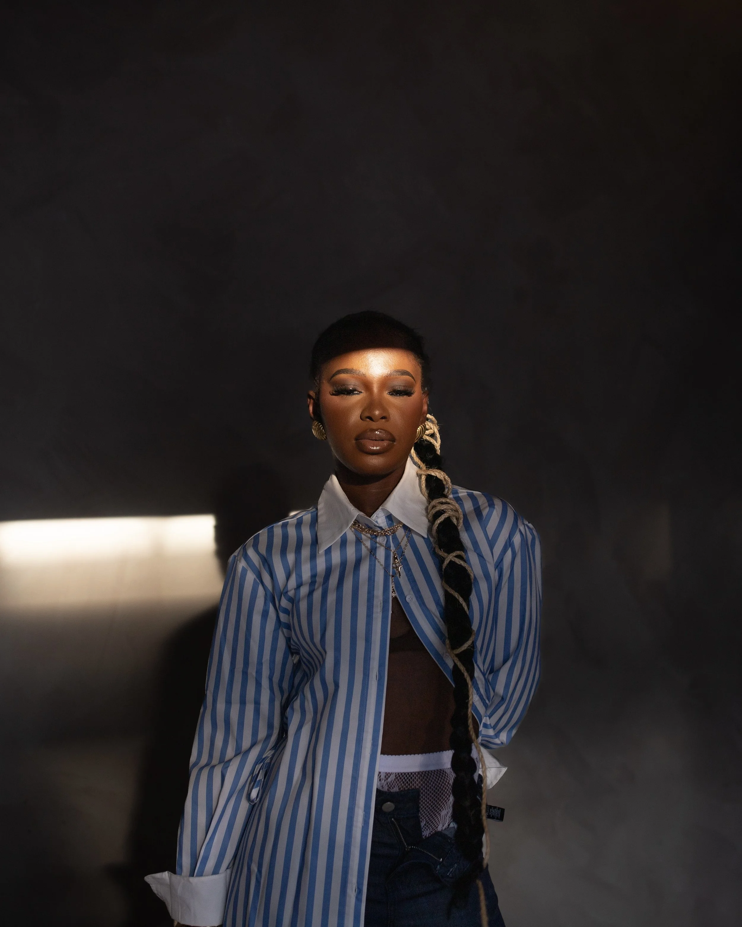 A woman with dark skin and long braided hair posing in a dark room with a light spot on the wall behind her, wearing a blue and white striped shirt, jewelry, and makeup.