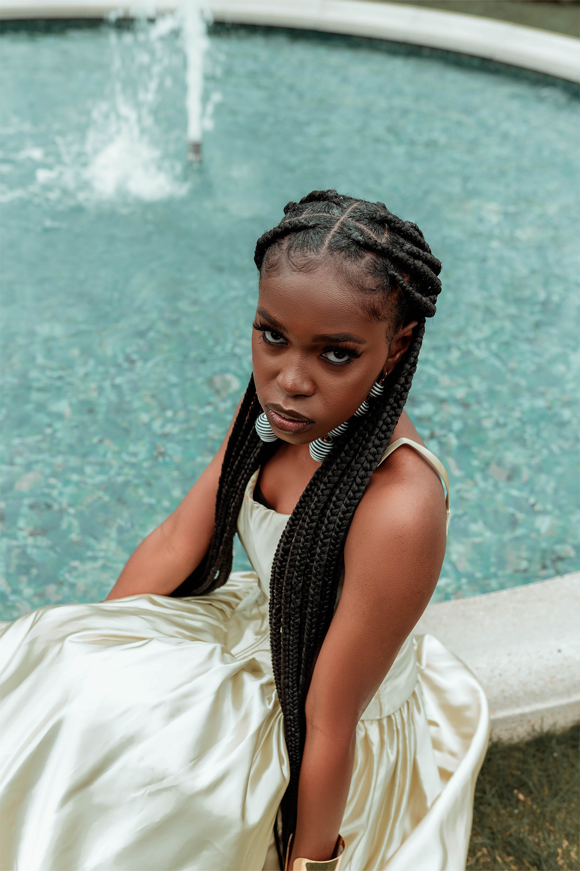 Queen Drie in a royal gown and a gold arm cuff, sitting by the fountain at the Bahai House of Worship in May 2022