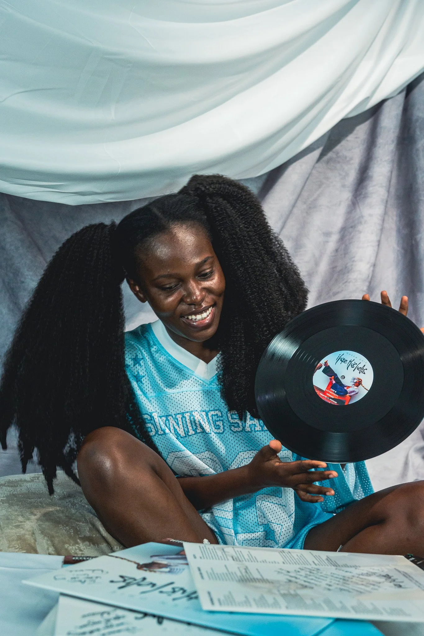 Queen Drie with long, black, textured hair, wearing a turquoise sports jersey, sitting on a couch, holding a vinyl record and smiling.