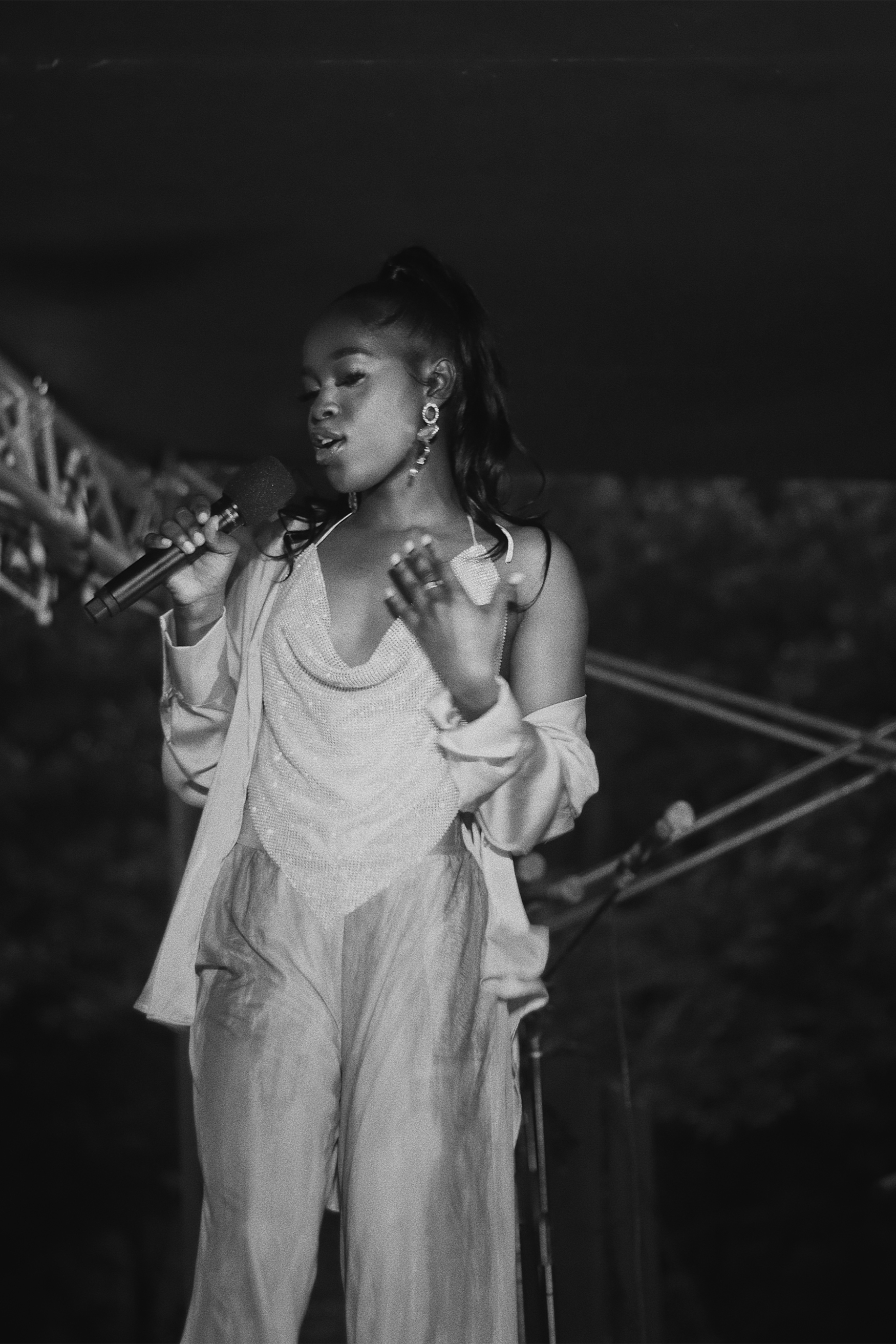 A Black woman singing into a microphone on stage at night, wearing elegant earrings and a sparkling dress with a light jacket.