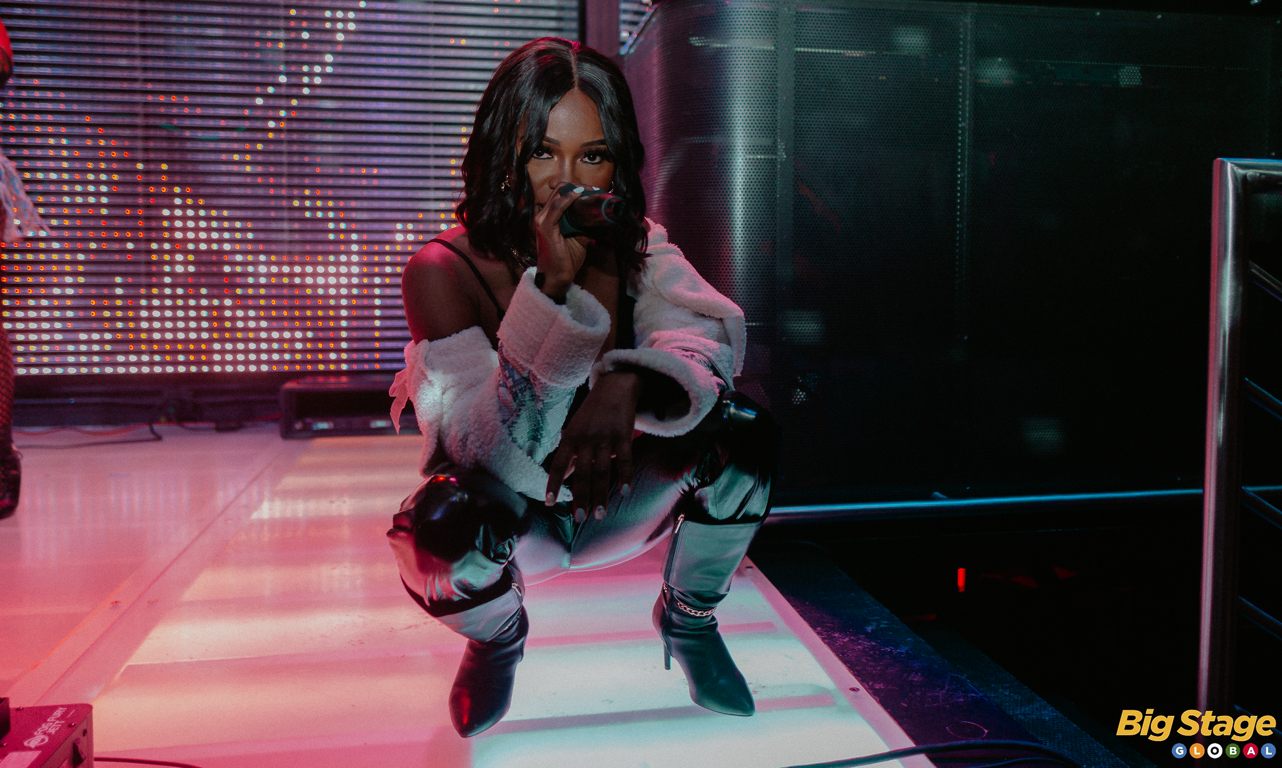 A woman performing on stage, squatting and holding a microphone near her mouth, with a colorful LED light backdrop and large black speakers behind her. The text 'Big Stage' is visible in the bottom right corner.