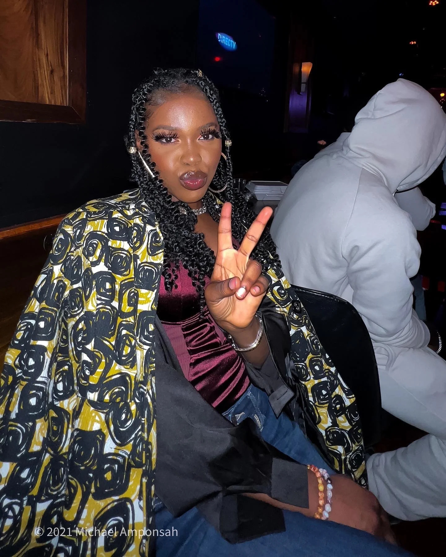 A woman with curly hair and makeup sitting in a dark room, making a peace sign with her right hand, wearing a patterned jacket and a bracelet.