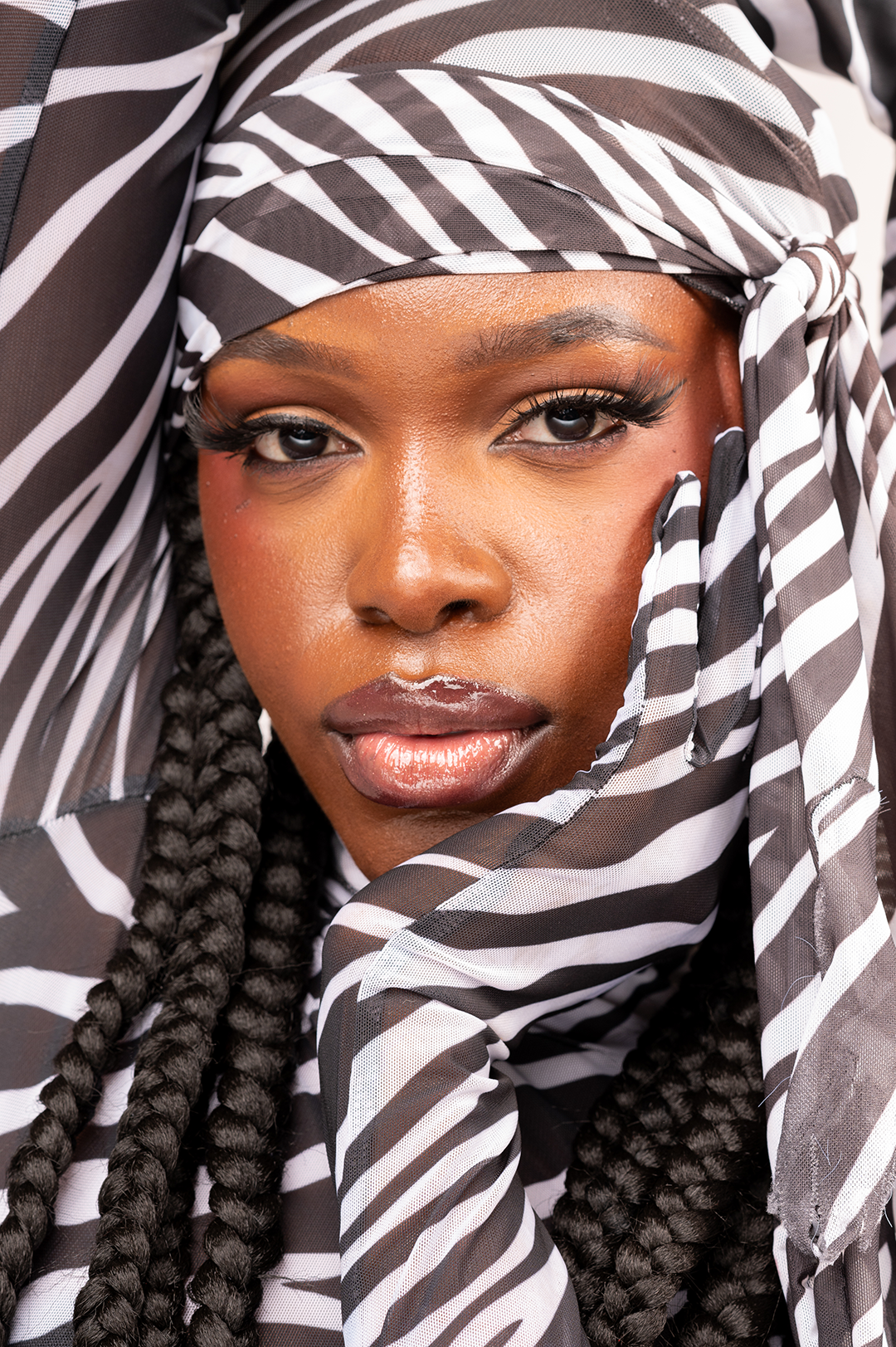 Close-up of Queen Drie with braided hair, wearing a zebra-striped headscarf, looking directly at the camera.