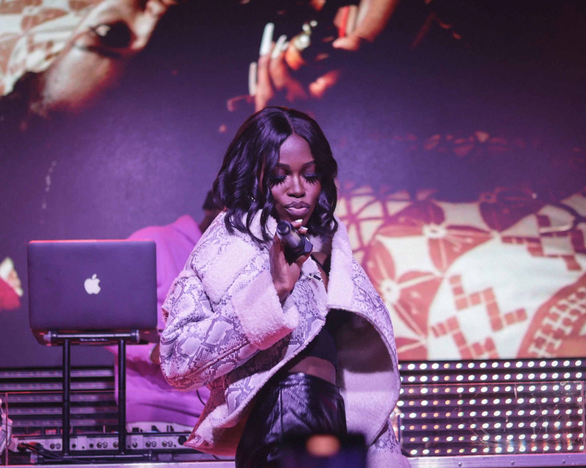 Queen Drie with dark, wavy hair, dressed in a patterned coat and black top, holding a microphone on stage with purple and pink lighting, with a DJ and equipment in the background at Stereo Nightclub.