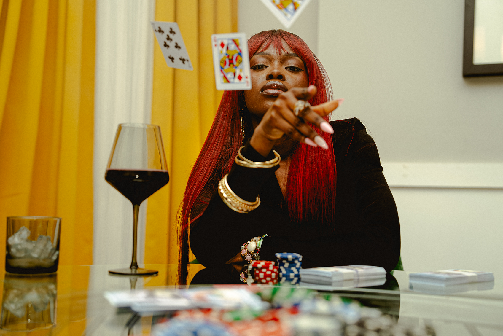 Queen Drie with red hair playing poker at a table with chips and cards, holding a playing card.