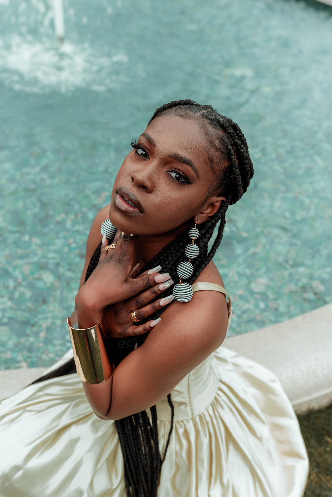 Queen Drie in a royal gown and a gold arm cuff, sitting by the fountain at the Bahai House of Worship in May 2022