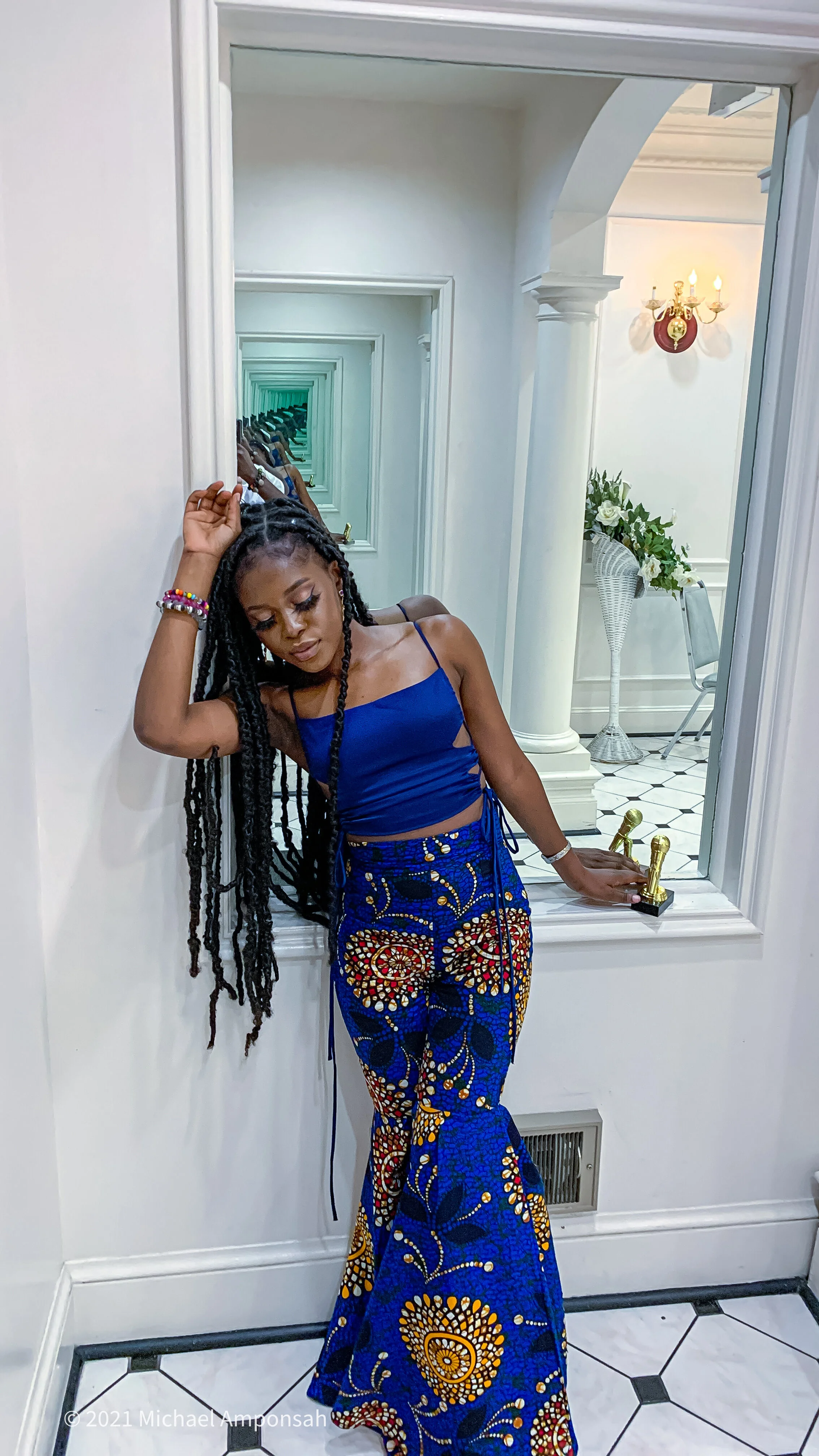 A young woman with long twisted hair wearing a blue crop top and matching patterned pants, leaning against a white wall with her eyes closed.