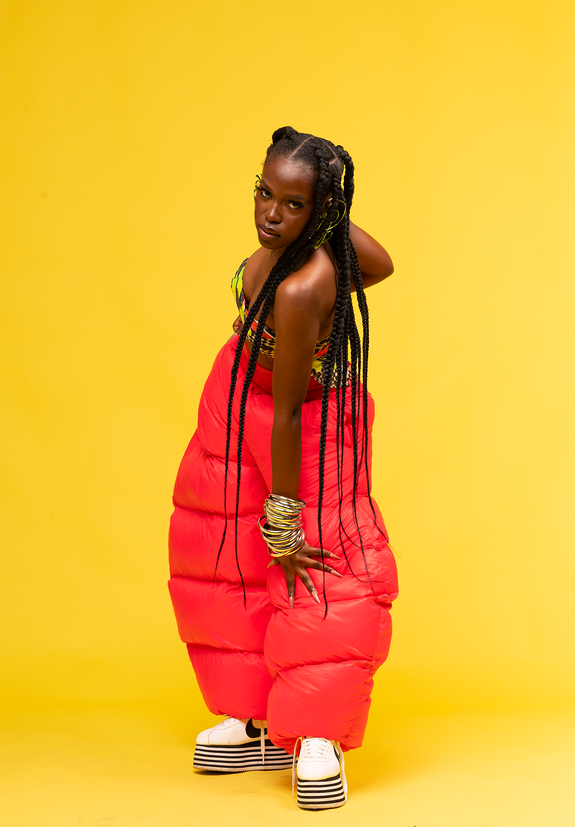Queen Drie with long, twisted hair wearing a colorful top, bright red puffer pants, and striped platform shoes, posing against a solid yellow background.