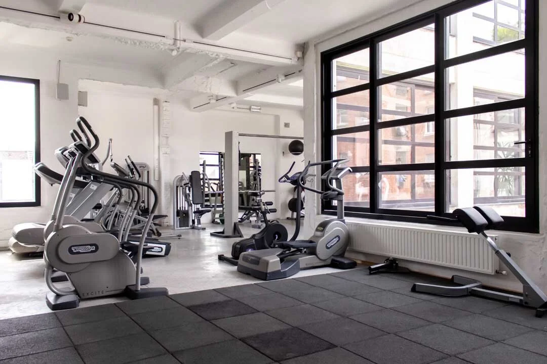 Empty gym with exercise machines including ellipticals, a rowing machine, and a weight machine, with large windows and black rubber mats on the floor.