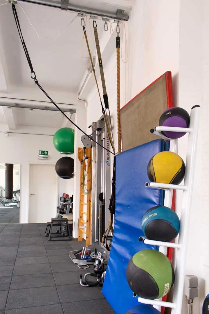 Gym area with fitness equipment including medicine balls on a rack, a punching bag, resistance bands hanging from the ceiling, a blue exercise mat, and other workout gear.
