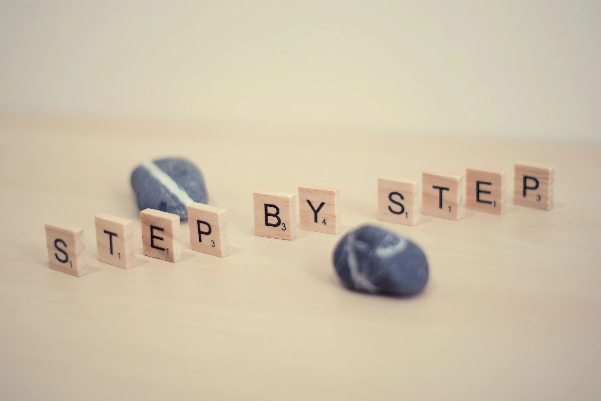 Scrabble tiles spelling 'STEP BY STEP' on a light wooden surface, with two small rocks placed near the tiles. Reminder to take your business growth ideas and activities one step at a time.