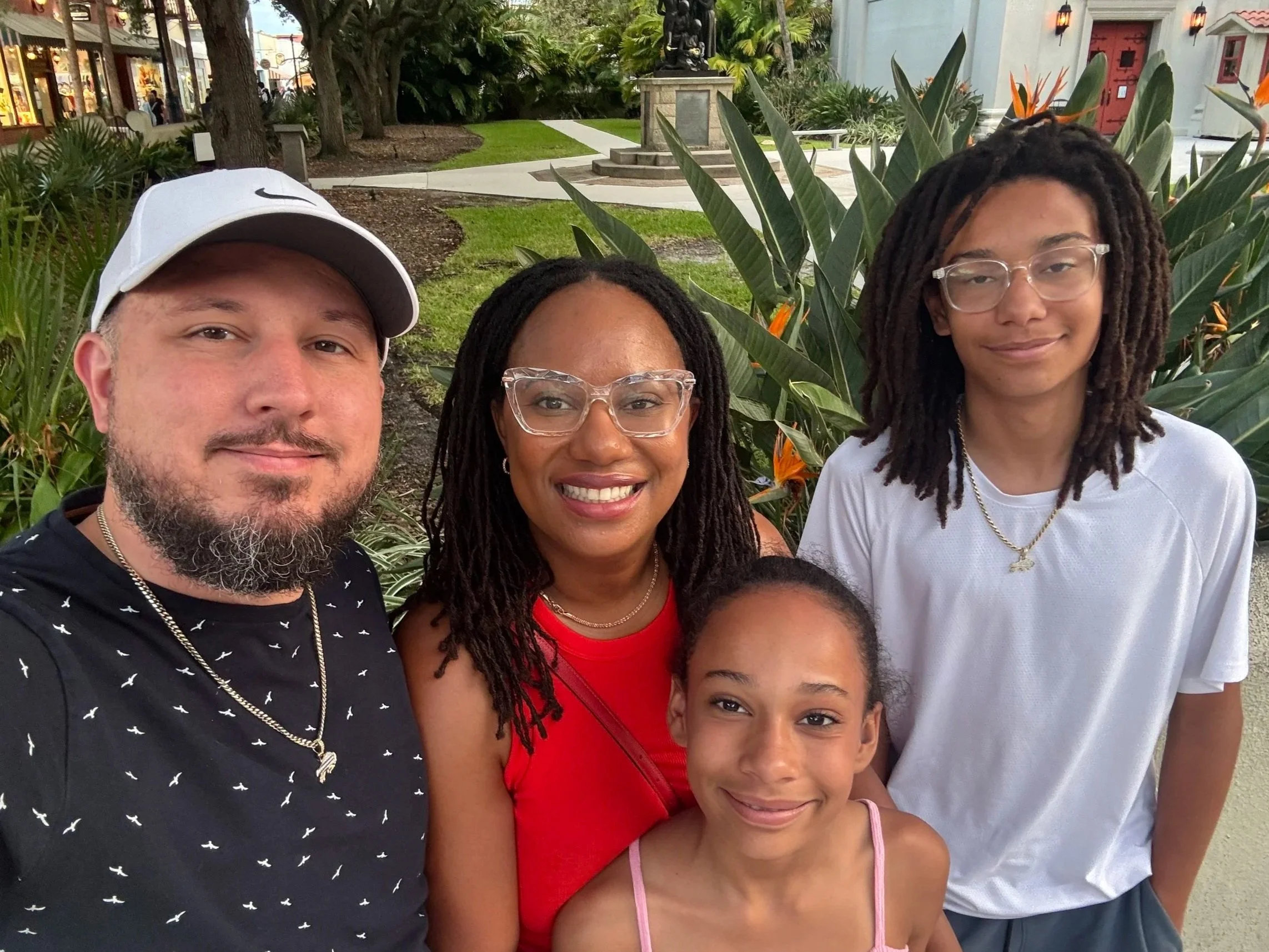 A happy family smiling for a photo outdoors. The group includes a husband and wife business team and their two children. They are standing in front of large green plants and a building with red doors and outdoor lighting.