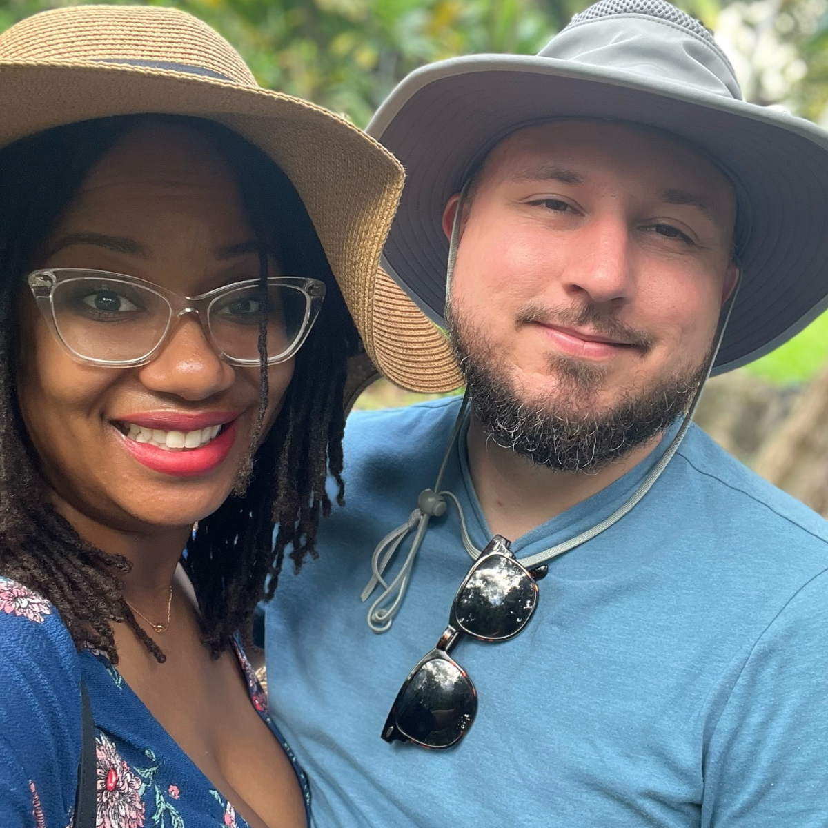A smiling couple in business together wearing wide-brimmed hats outdoors while on vacation together, with a woman wearing glasses and a colorful dress, and a man with sunglasses hanging from his shirt collar, surrounded by greenery.