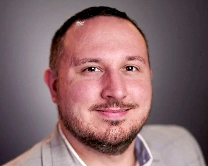 A professional portrait / headshot of a businessman with short hair, a goatee, and a light-colored suit. He is smiling and has a neutral gray background.