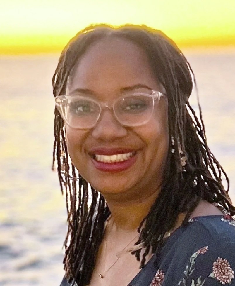 A black woman with braided hair wearing glasses and a floral dress smiling at the camera with a body of water and sunset in the background.