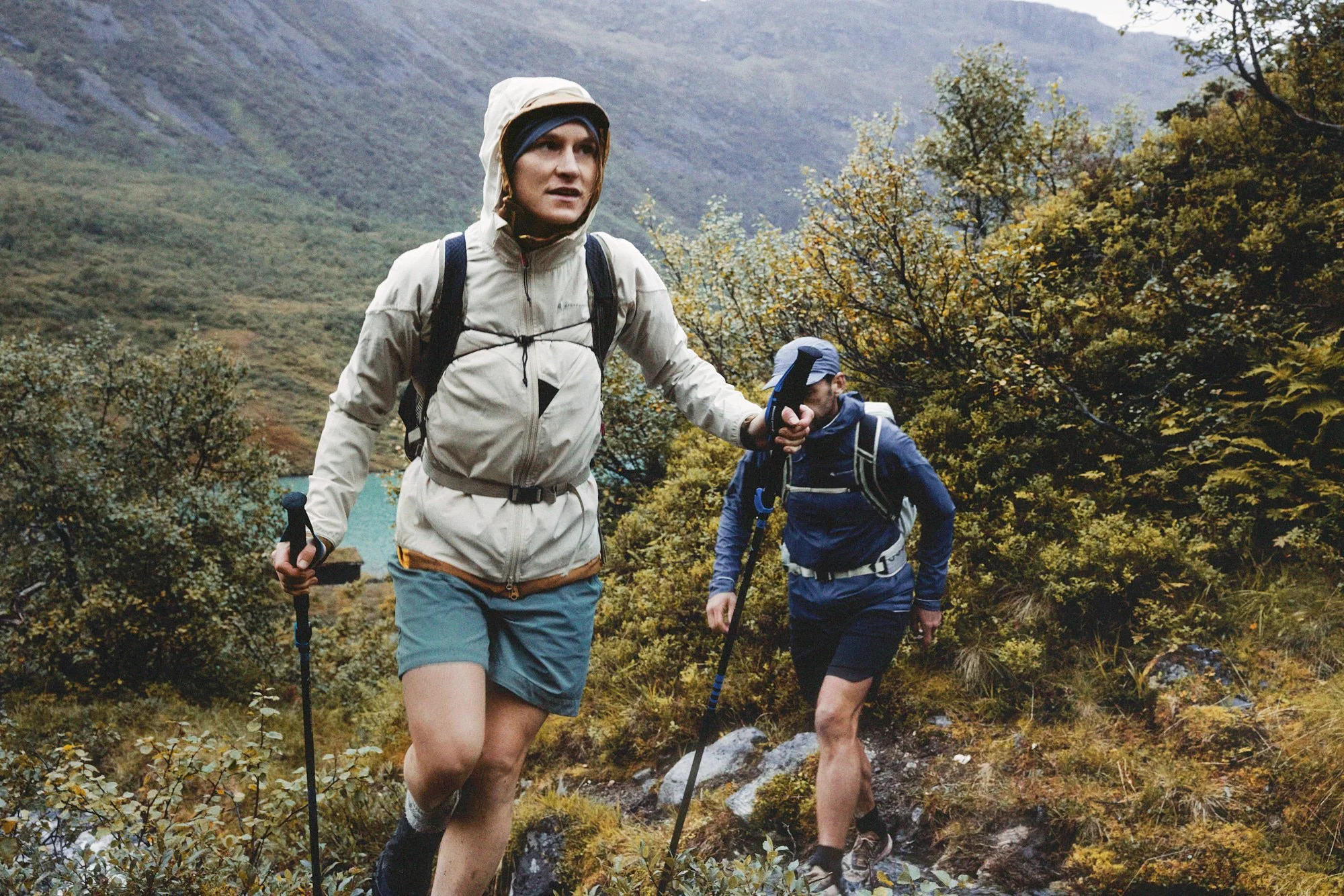 Two hikers trekking through a lush, rugged mountain trail surrounded by dense autumn foliage.