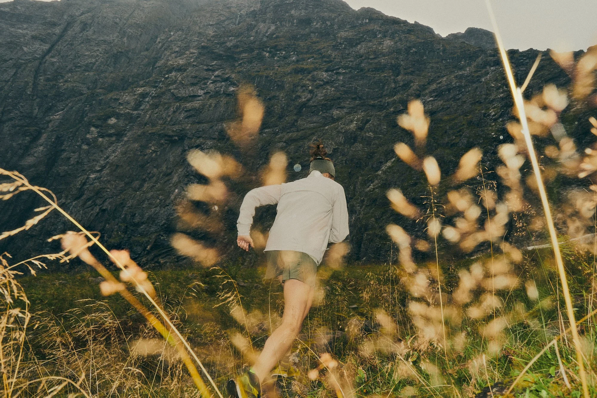 A person running up a grassy slope next to a large black rock formation, with tall grass and plants in the foreground.