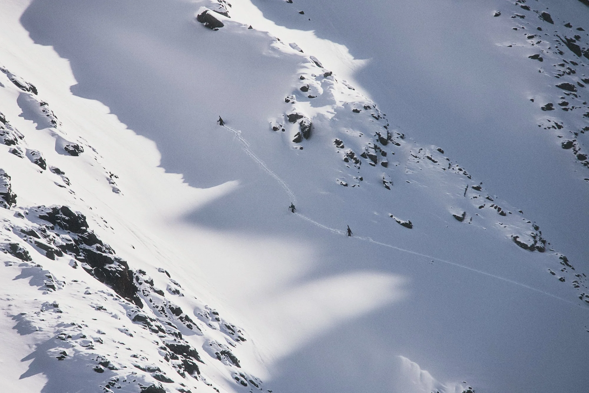 A snowy mountain slope with several skiers making their way down, leaving tracks in the snow, surrounded by rocks and snow-covered terrain.