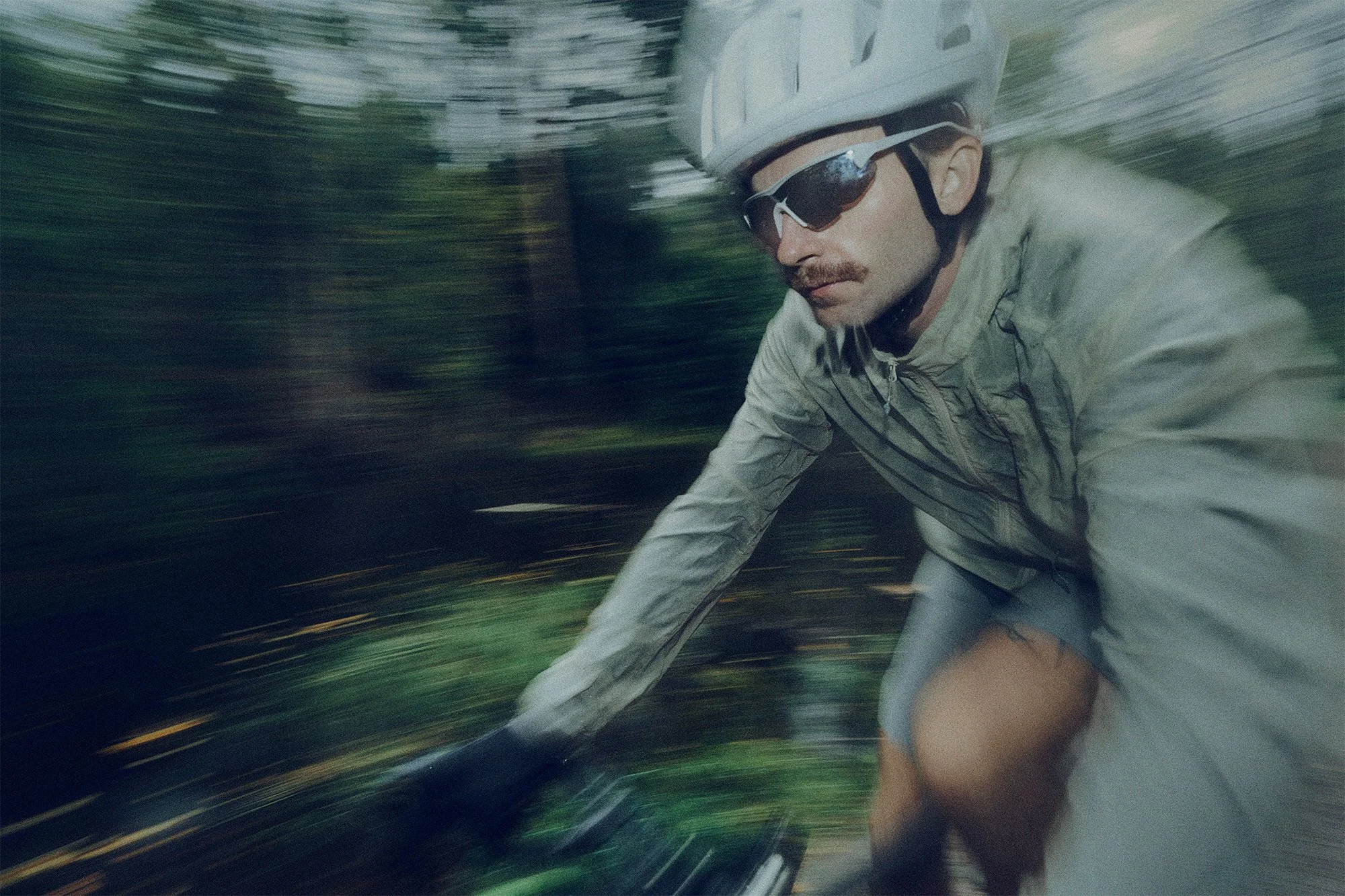 A man riding a bicycle in motion wearing a gray helmet, sunglasses, and a light gray jacket with a blurred background of trees.