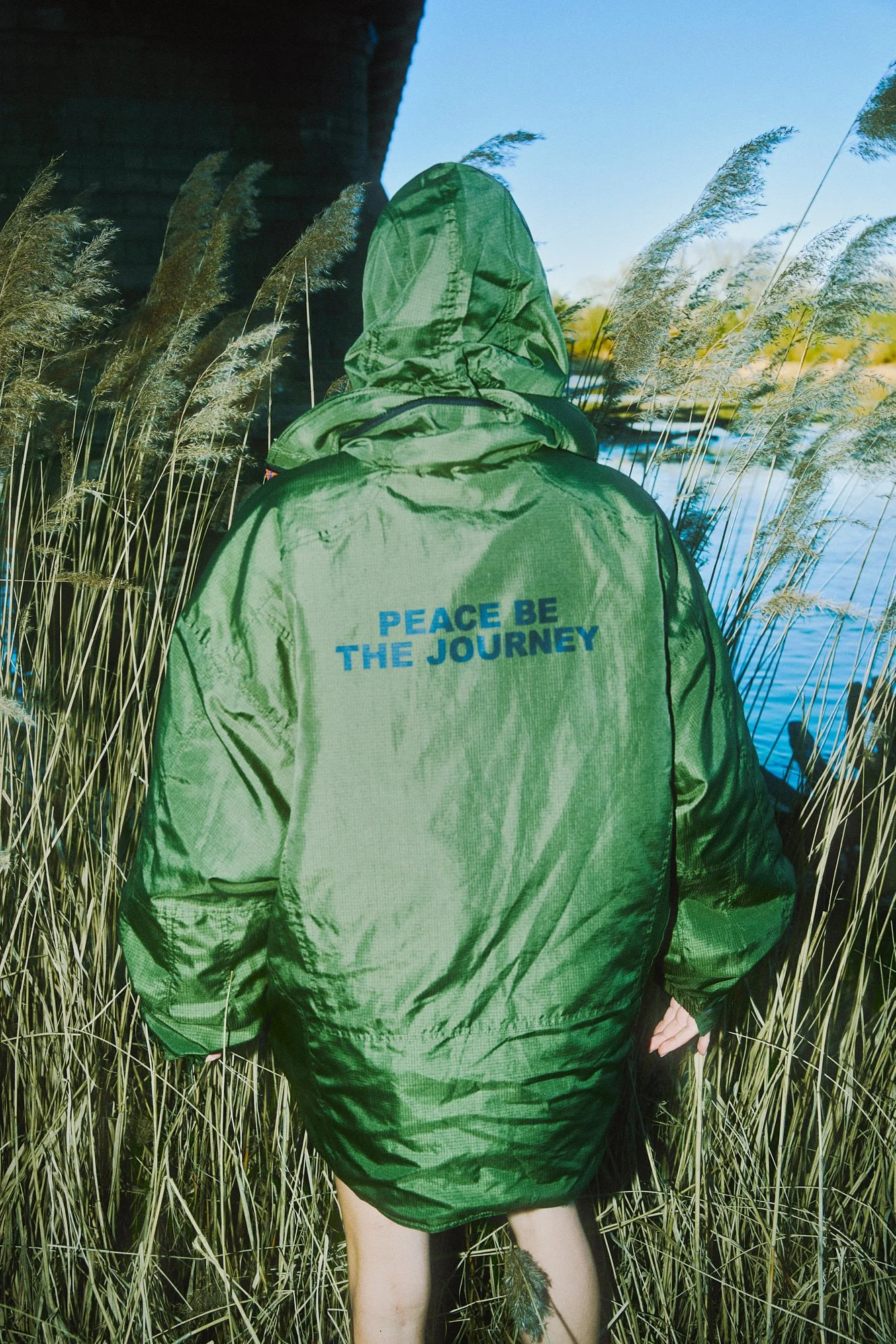 Person with a hooded green jacket with the words "PEACE BE THE JOURNEY" on the back, standing among tall grass near a body of water, with a bridge overhead.