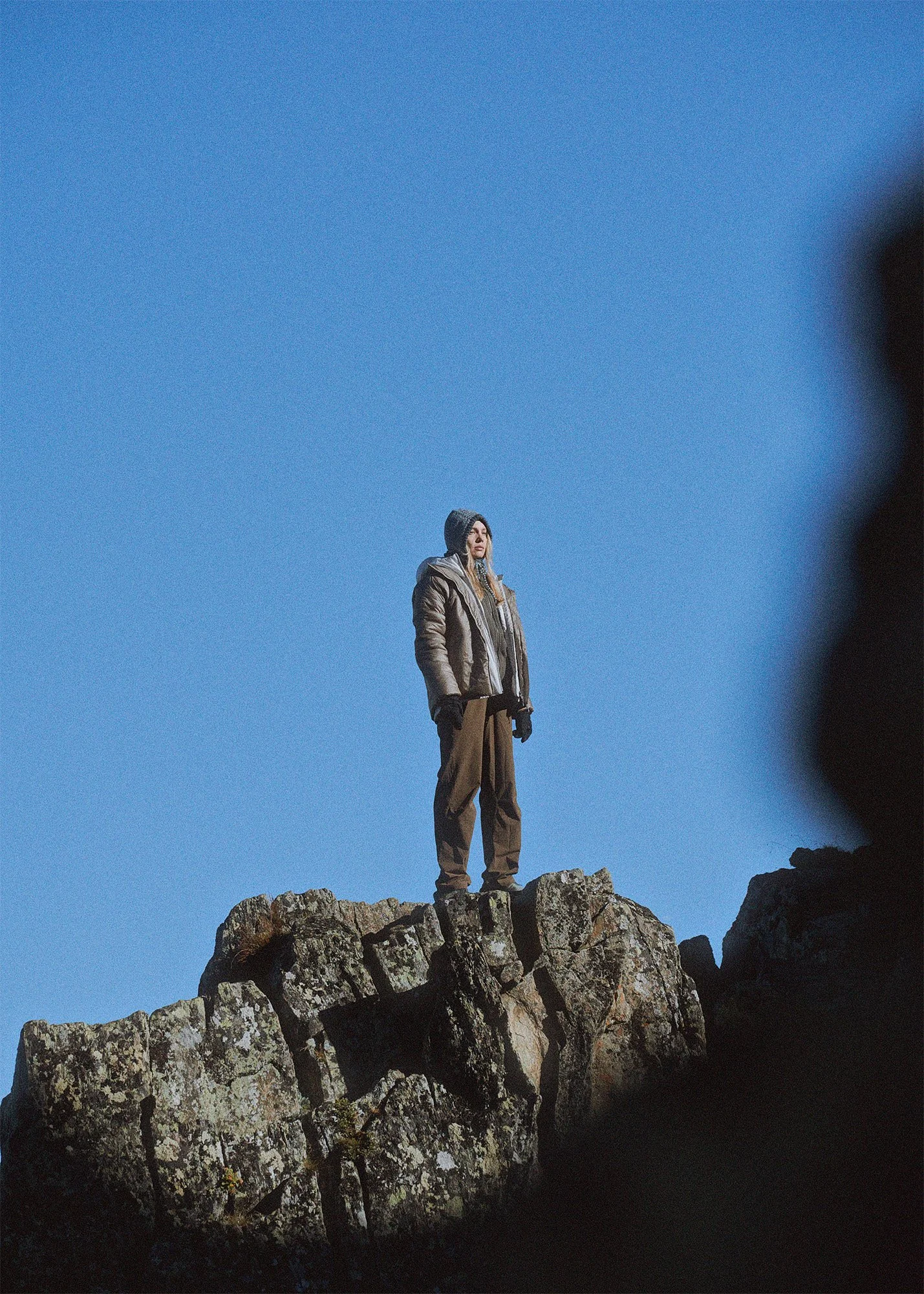 A woman dressed in outdoor winter clothing, standing on large rocks against a clear blue sky.