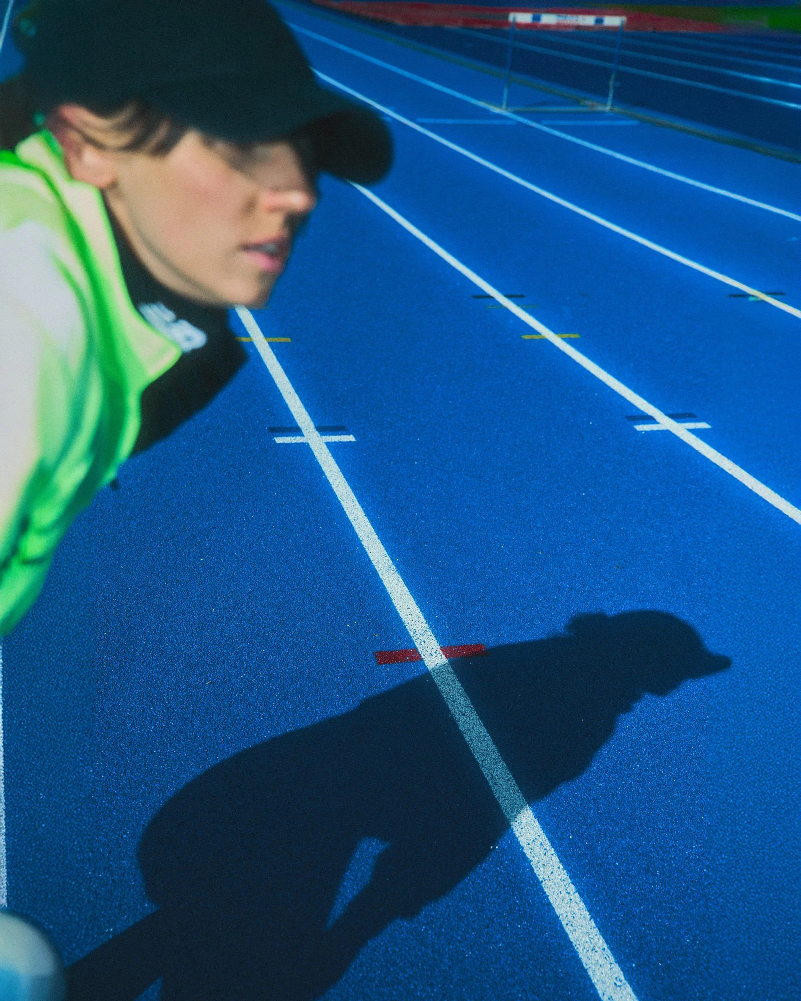 A female runner in a neon green jacket and black cap preparing on a blue outdoor track, with her shadow cast on the ground.