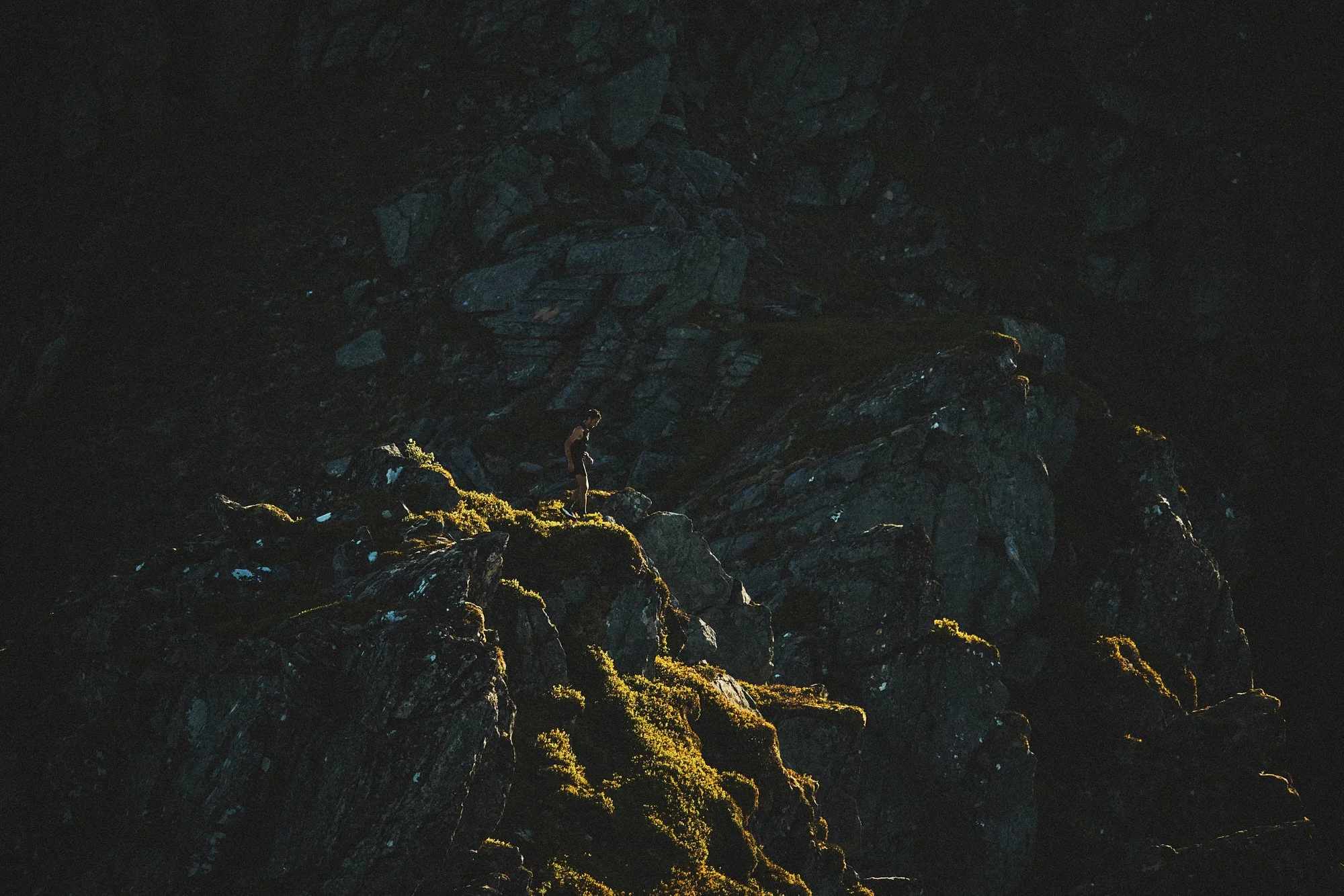 A person standing on moss-covered rocks at the edge of a dark, shadowy rocky cliff or mountain.