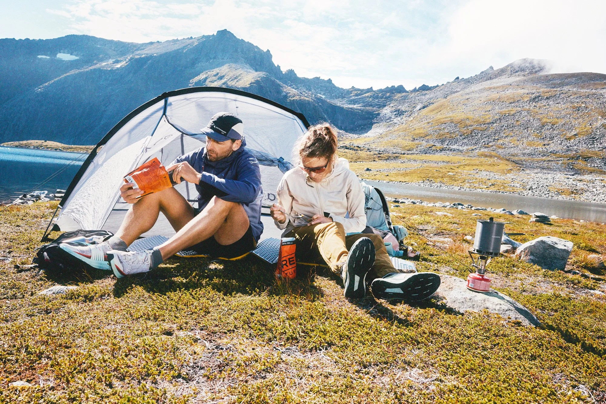 Two people sitting outdoors near a camping tent with mountain scenery in the background, camping stove on a rock, and books or maps in their hands.