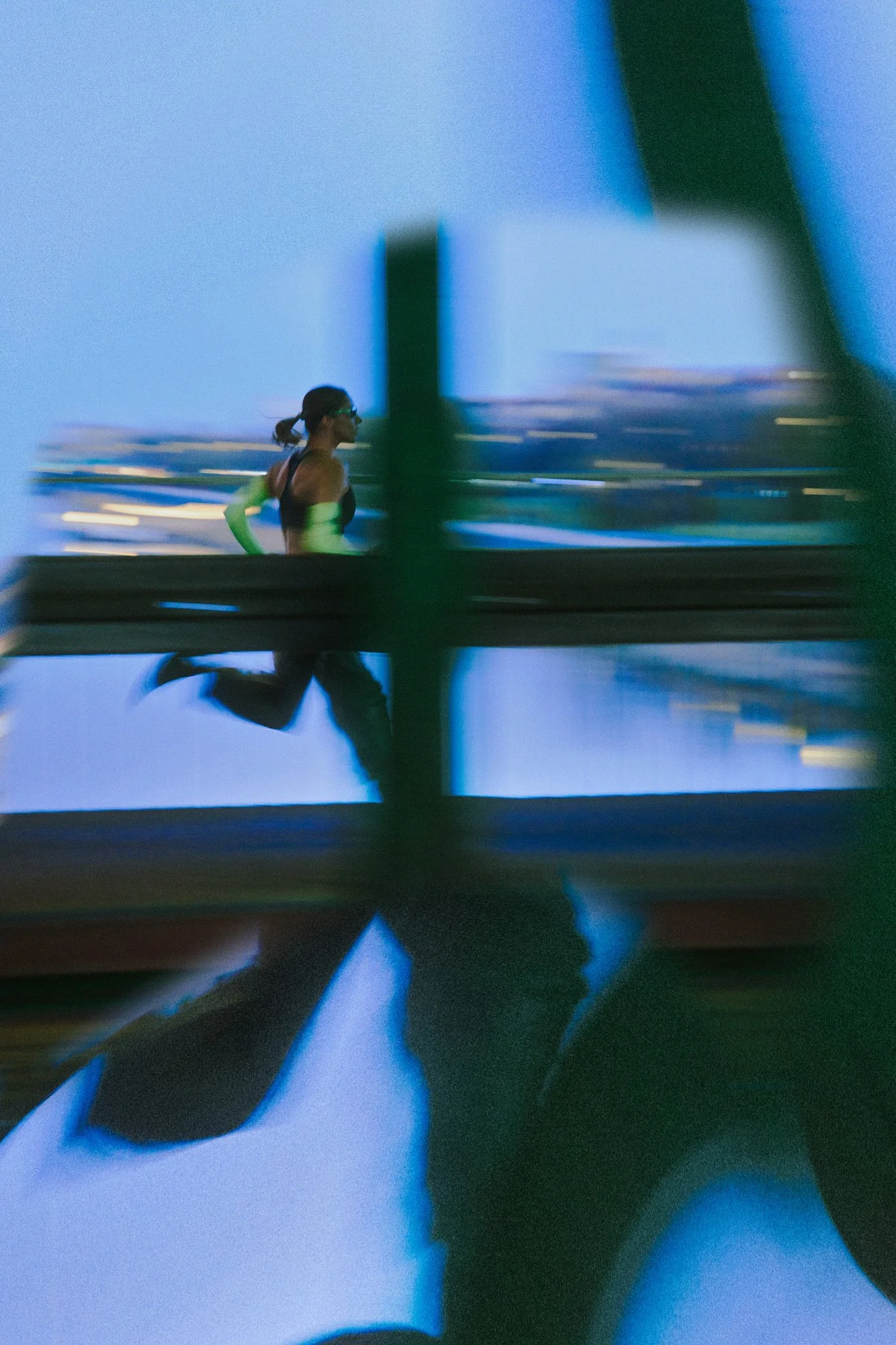 A person running on a bridge with a city skyline in the background during dusk, seen through a blurred window or structure.